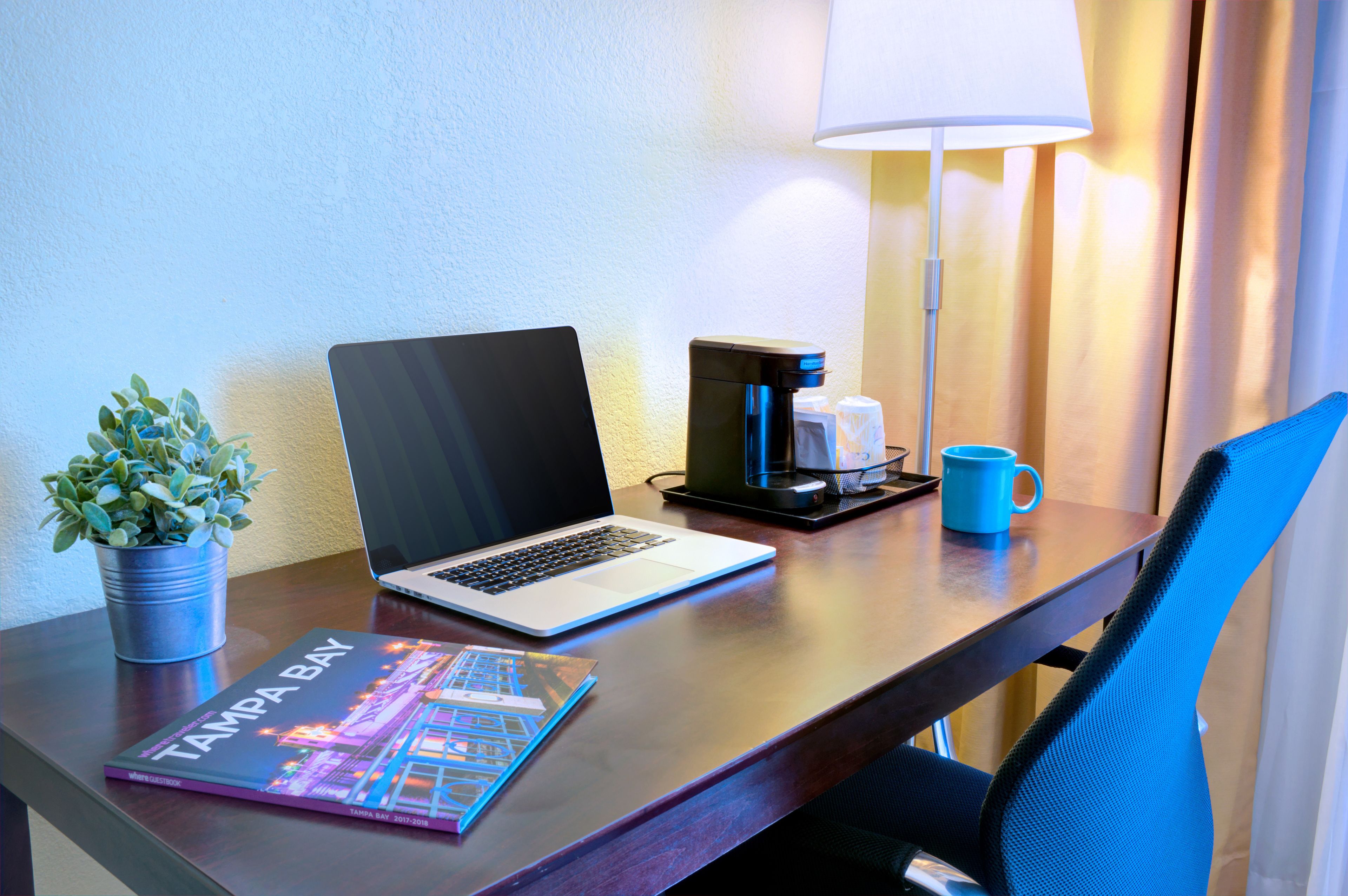 A hotel desk with a laptop, a coffee maker, a mug, a plant, and a book titled "Tampa Bay."