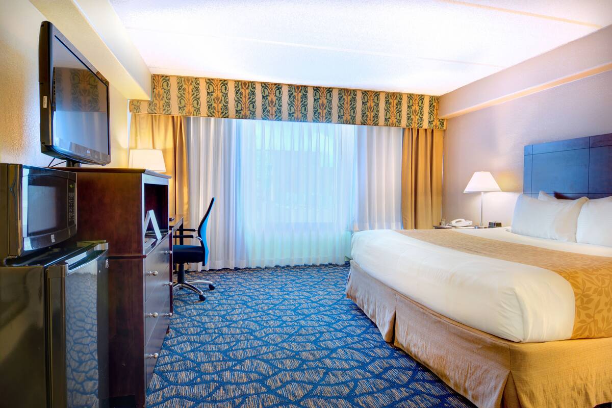 A hotel room with a large bed, a desk with a computer, a television, and a window with curtains.