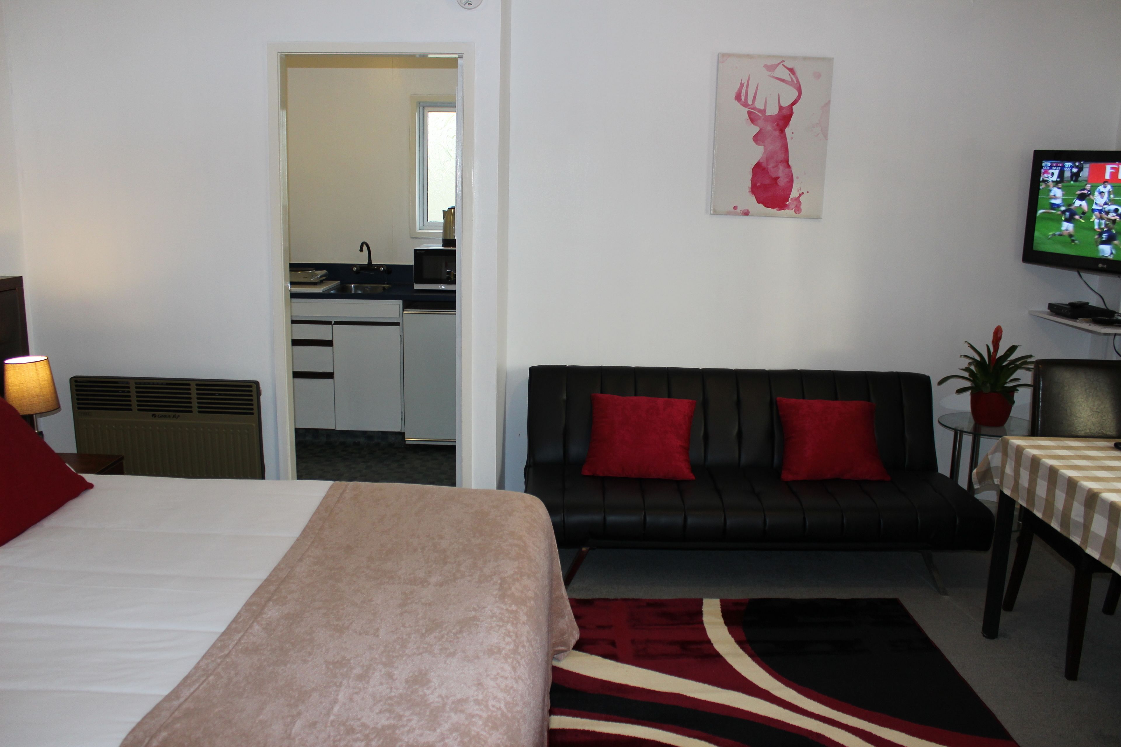 A hotel room with a bed, a black sofa with red cushions, a television, and a small table with a checkered tablecloth.