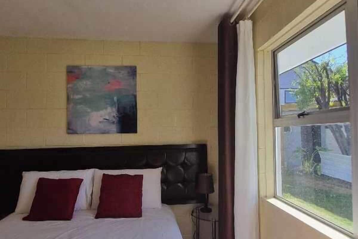 A hotel room with a bed, a nightstand, a lamp, a window with curtains, and a painting on the wall.
