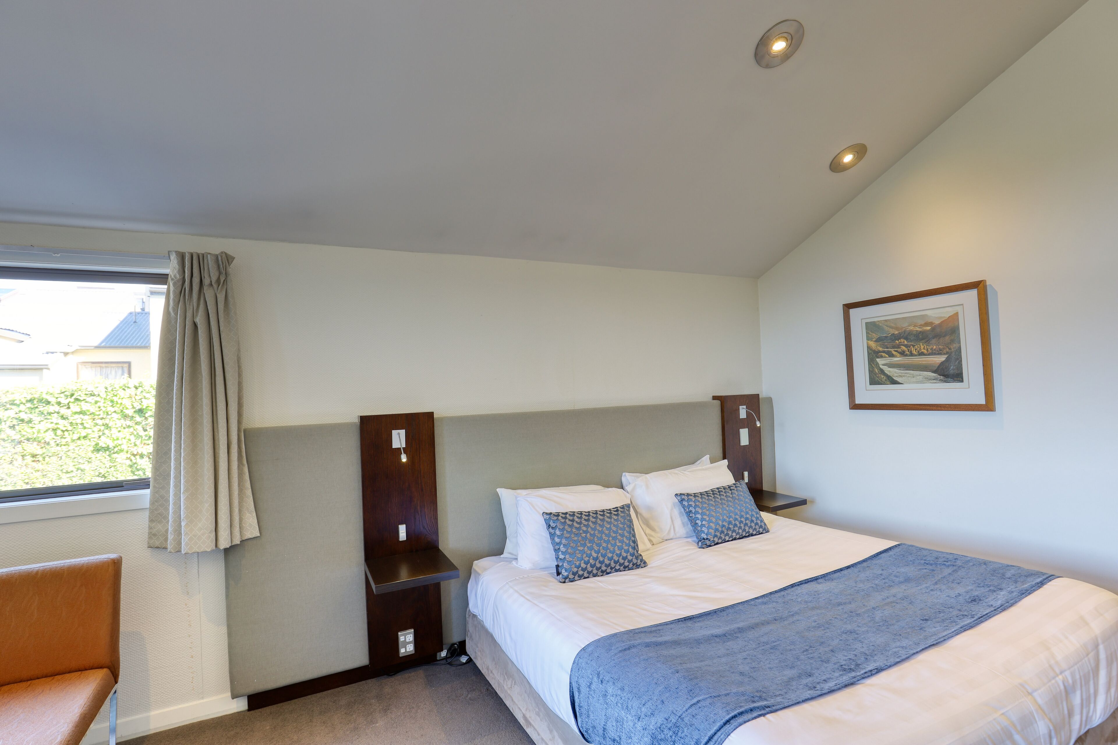 Une chambre à coucher avec un lit, une table de chevet, une chaise, une fenêtre avec des rideaux et un tableau encadré au mur.
