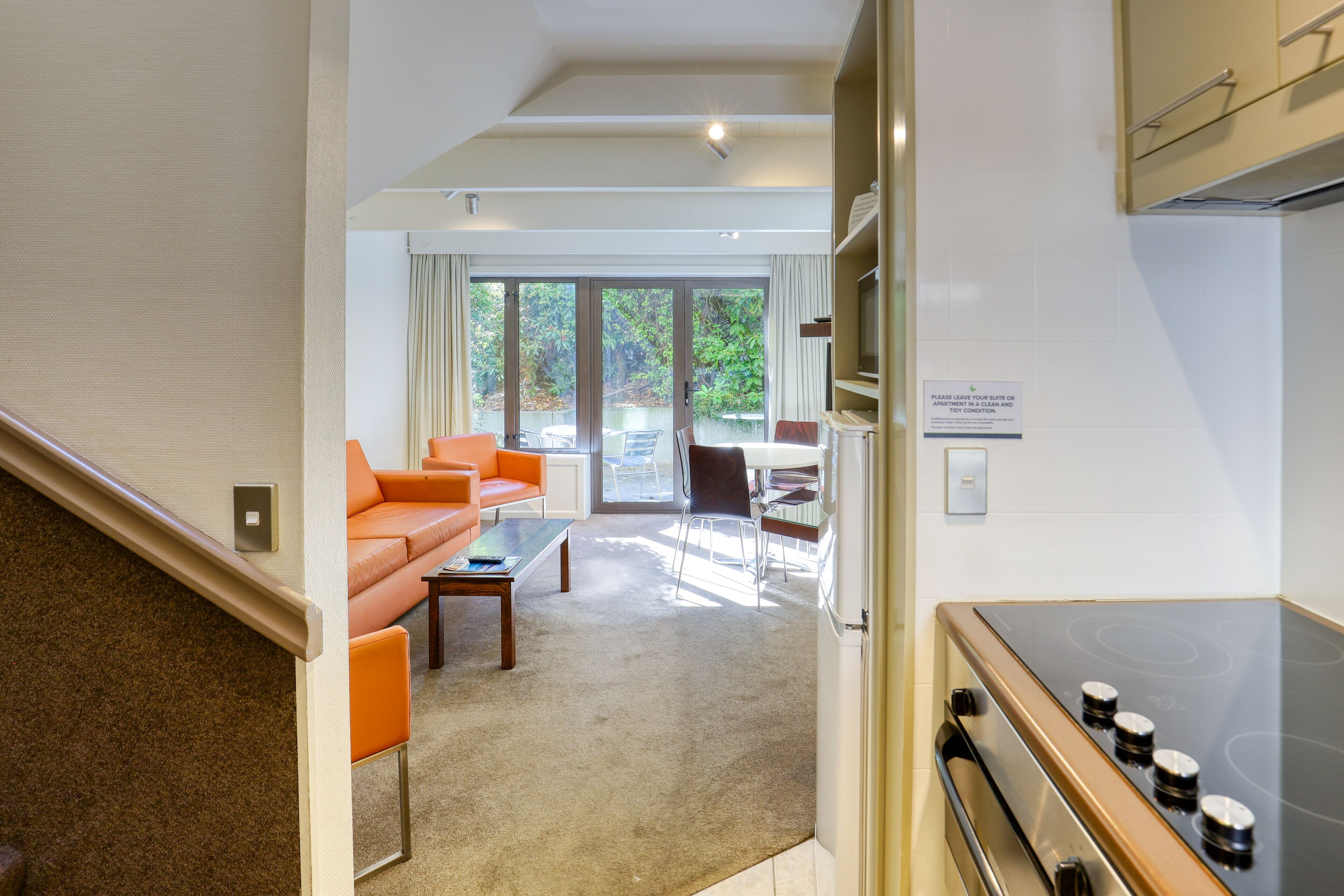 Una habitación de hotel moderna con zona de estar que incluye un sofá, sillas y una mesa de centro. Cuenta con un área de cocina con estufa y armarios. La habitación tiene un amplio ventanal corredero que da a un espacio exterior.
