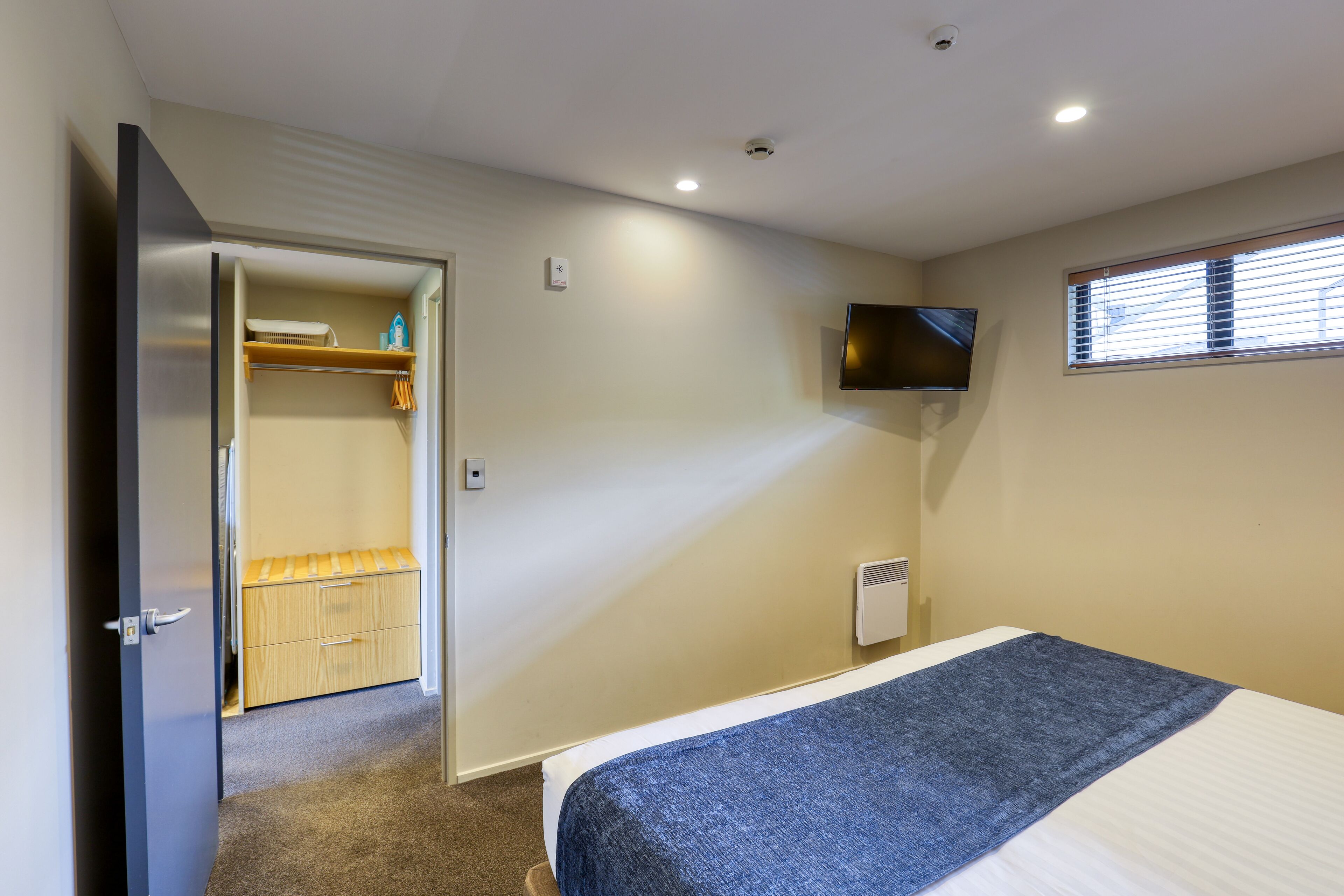 A bedroom with a bed, a TV mounted on the wall, a window with blinds, and a closet with a mirror.