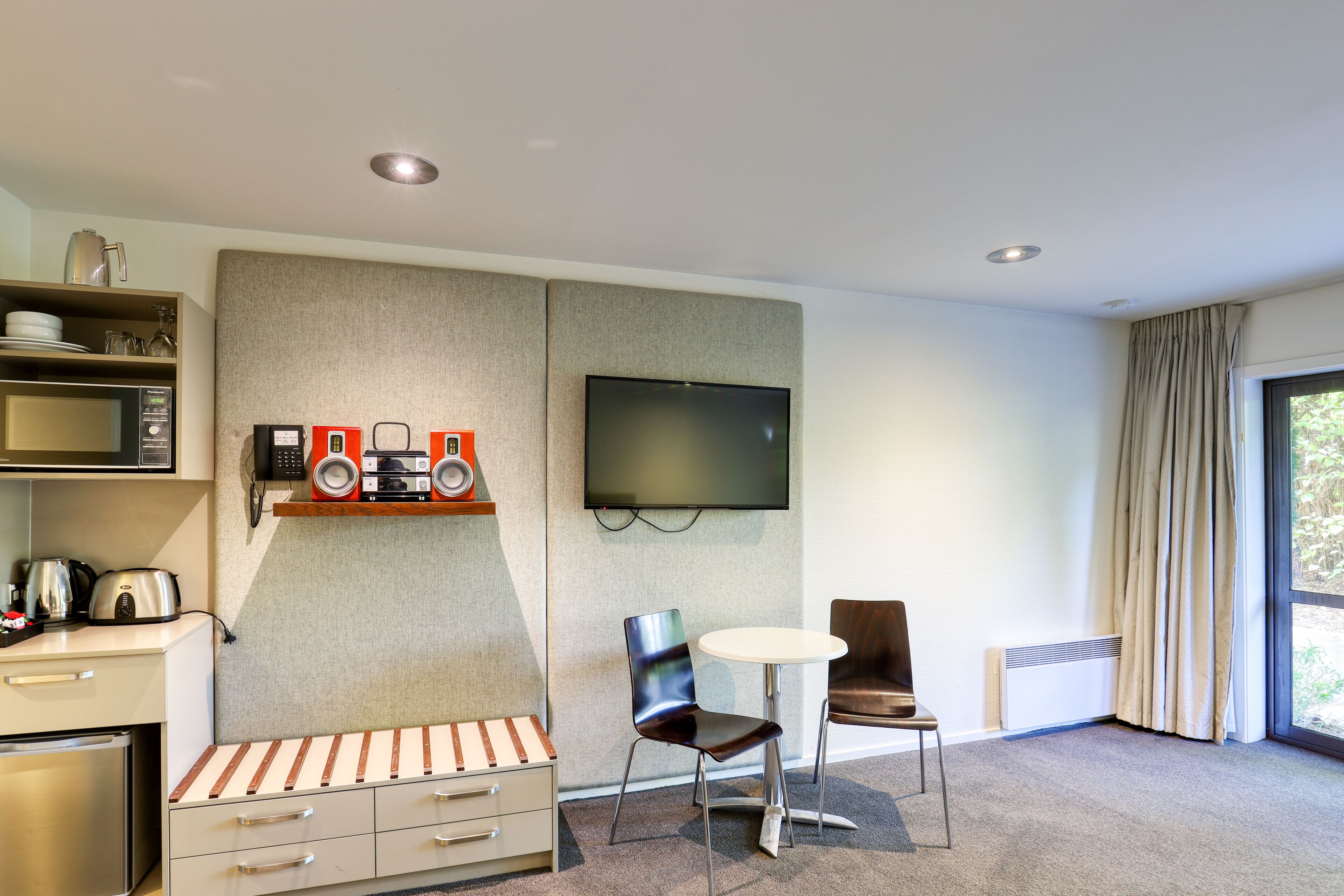 A modern hotel room with a kitchenette, a flat-screen TV, a round dining table, and a comfortable seating area.