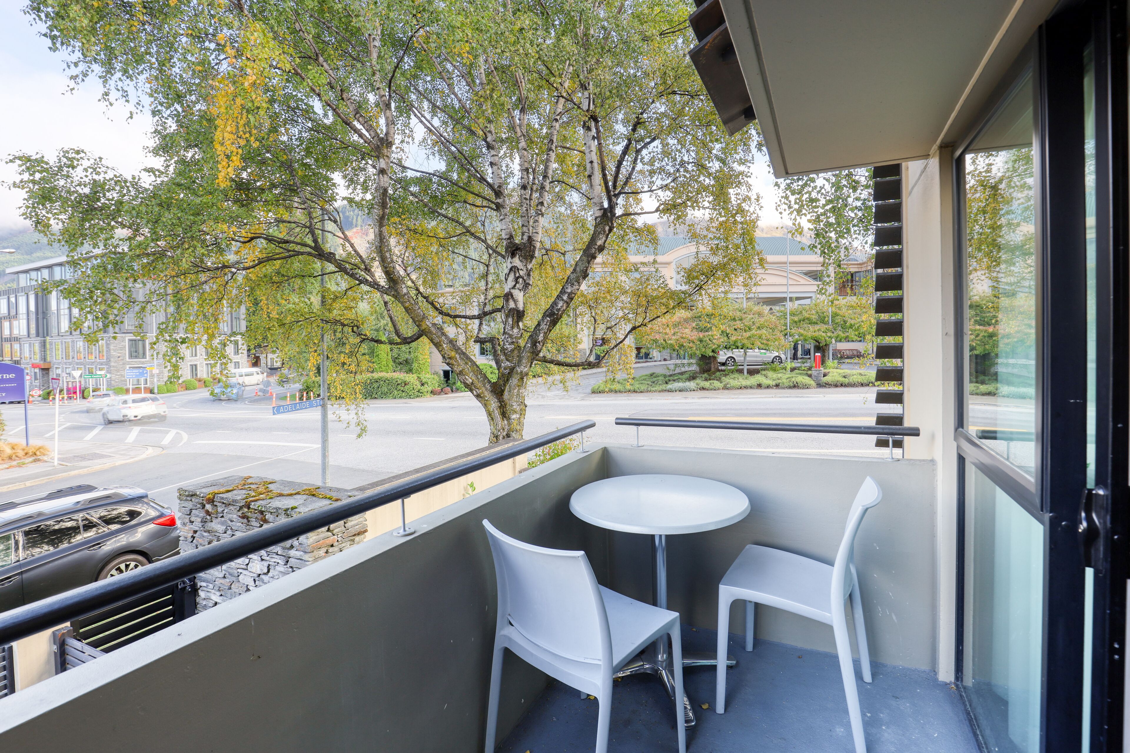 Un balcon avec une table et deux chaises, donnant sur une rue animée par la circulation des voitures et bordée d’immeubles.
