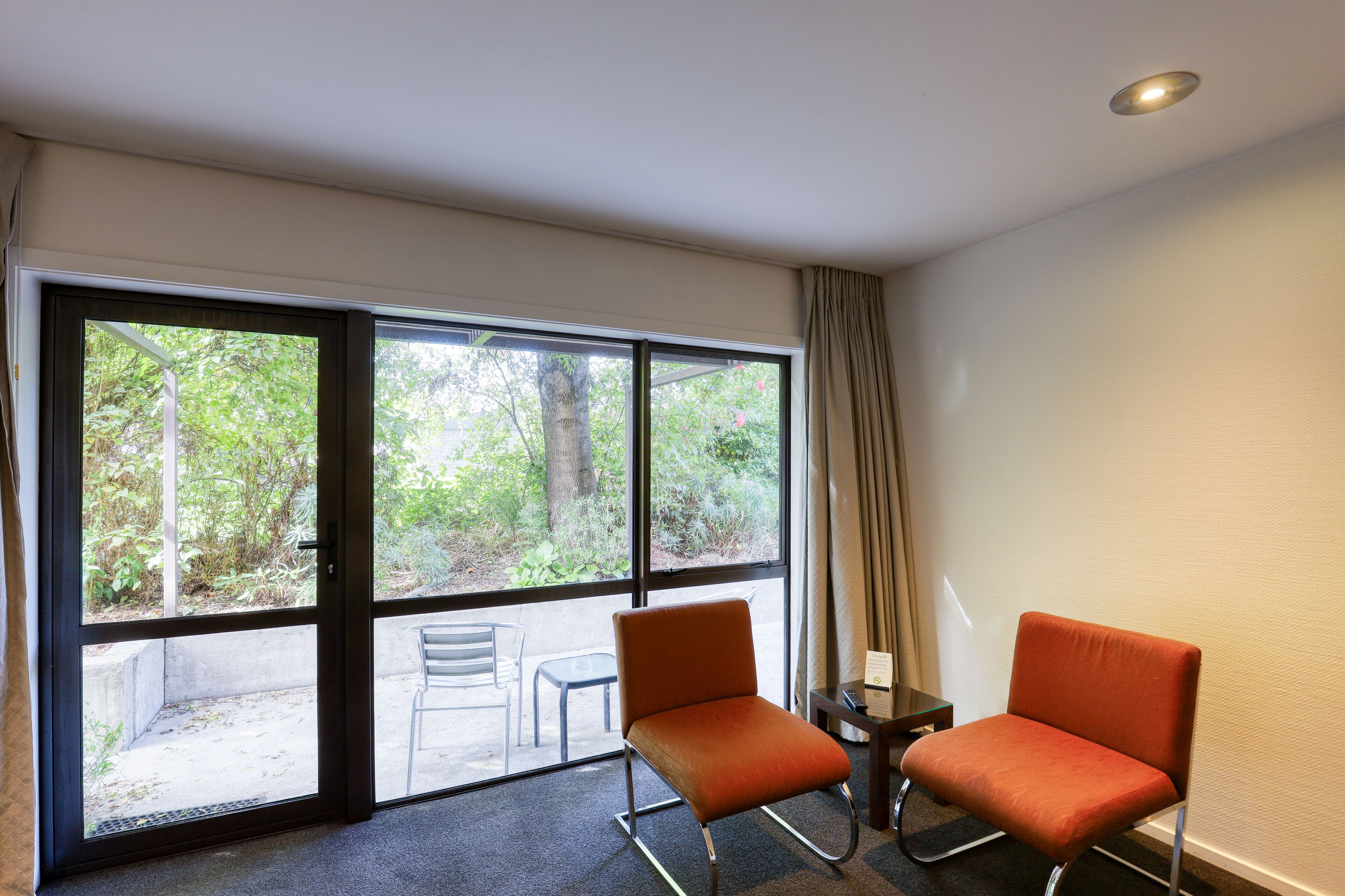 A room with two orange chairs, a small table, and a view of greenery through large windows.