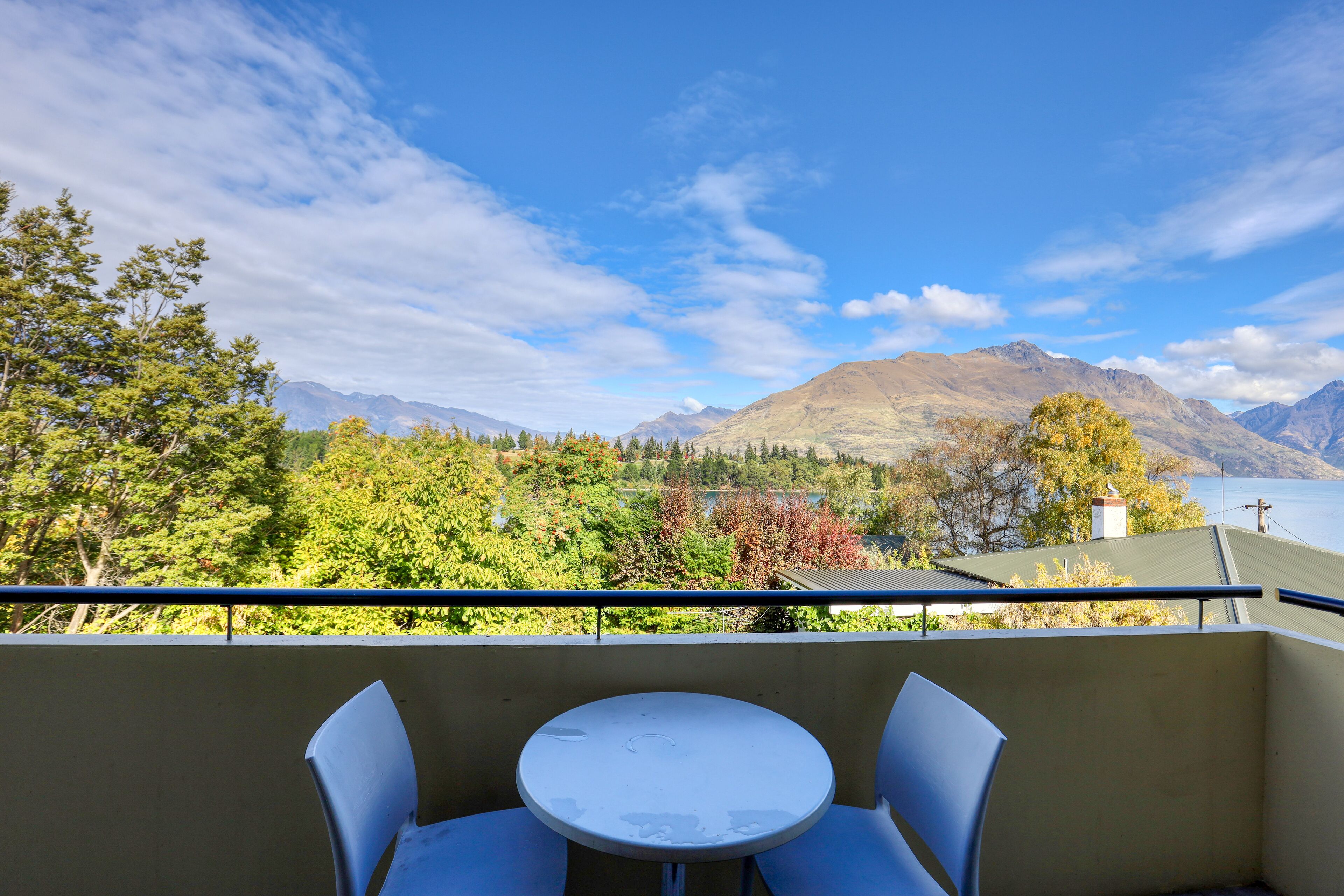 Un balcón con una mesa redonda y dos sillas, con vistas a un paisaje montañoso que incluye un lago y árboles.