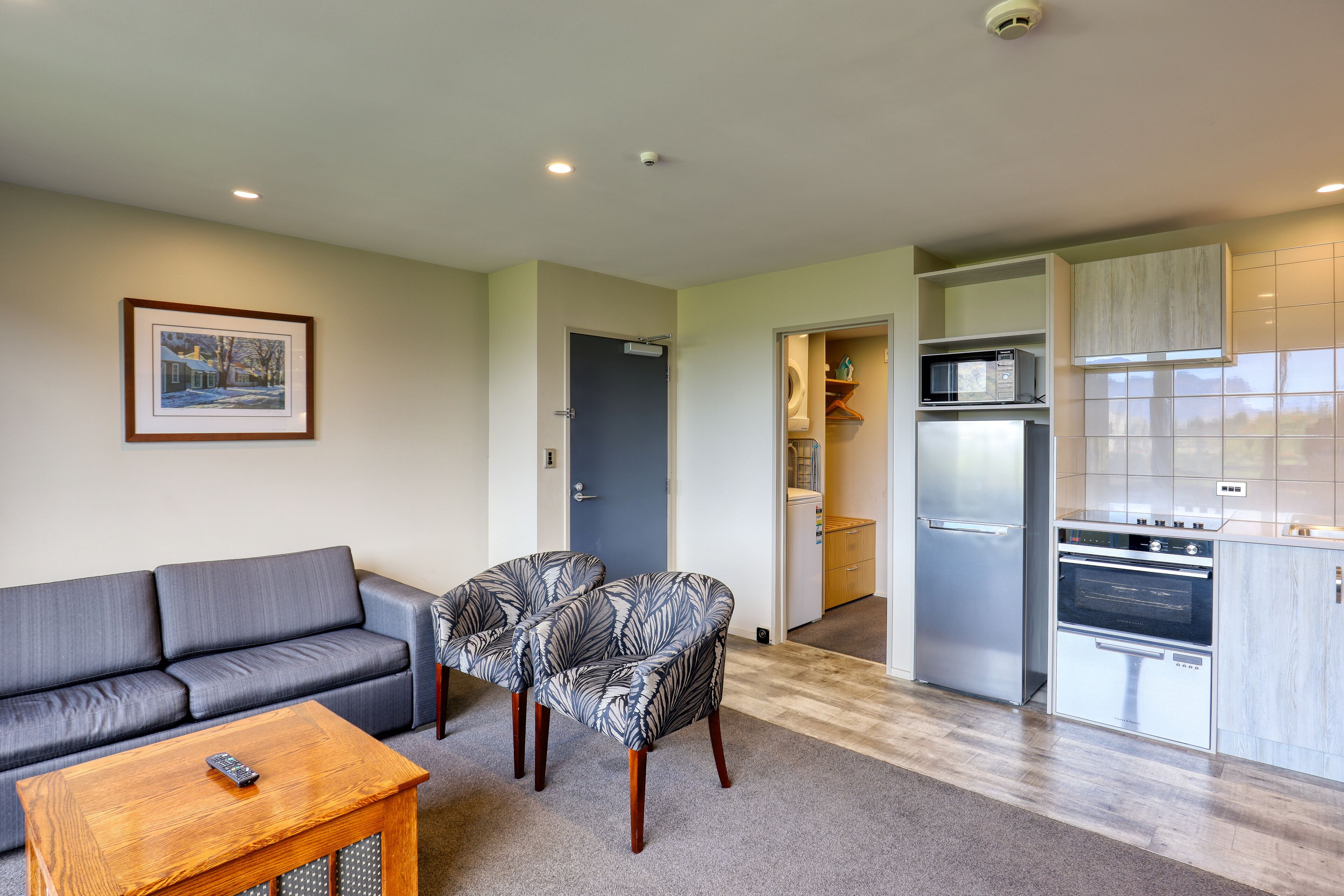 A living room with a gray sofa, patterned armchairs, and a wooden coffee table. A remote control is on the table. The kitchen has stainless steel appliances and light-colored cabinets.