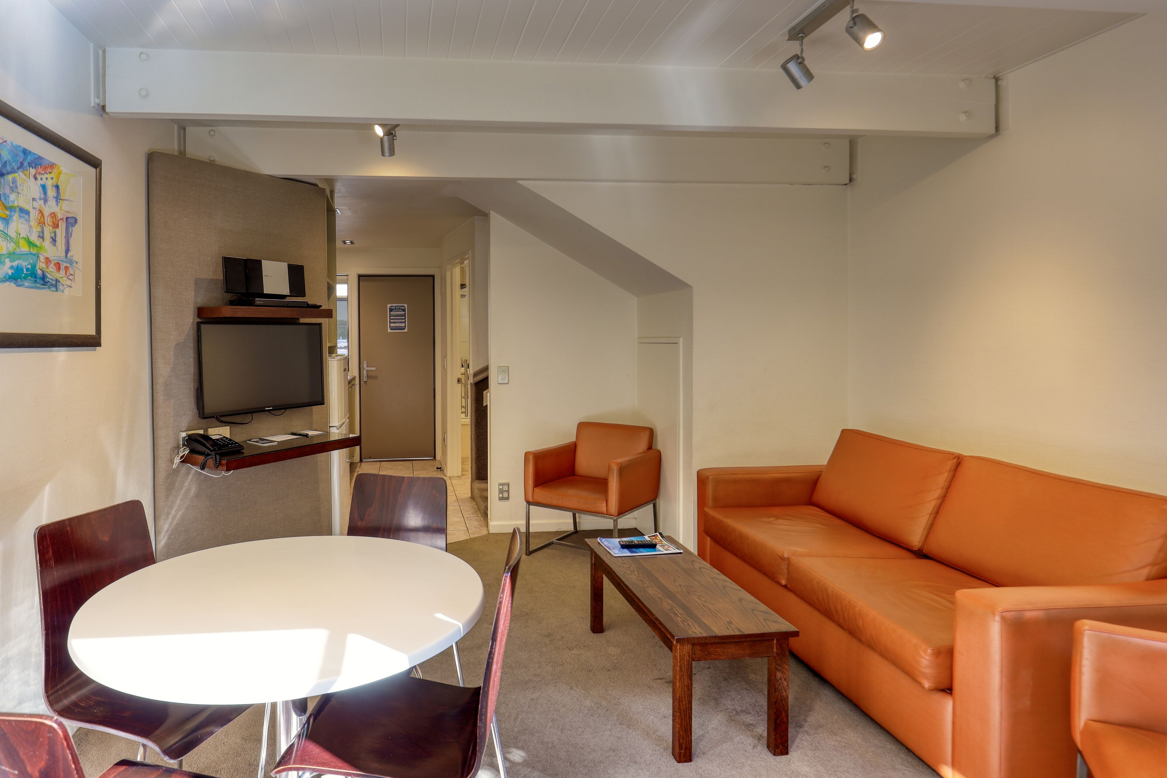 A modern living room with a white round table, brown leather chairs, an orange leather sofa, and a television on a shelf.
