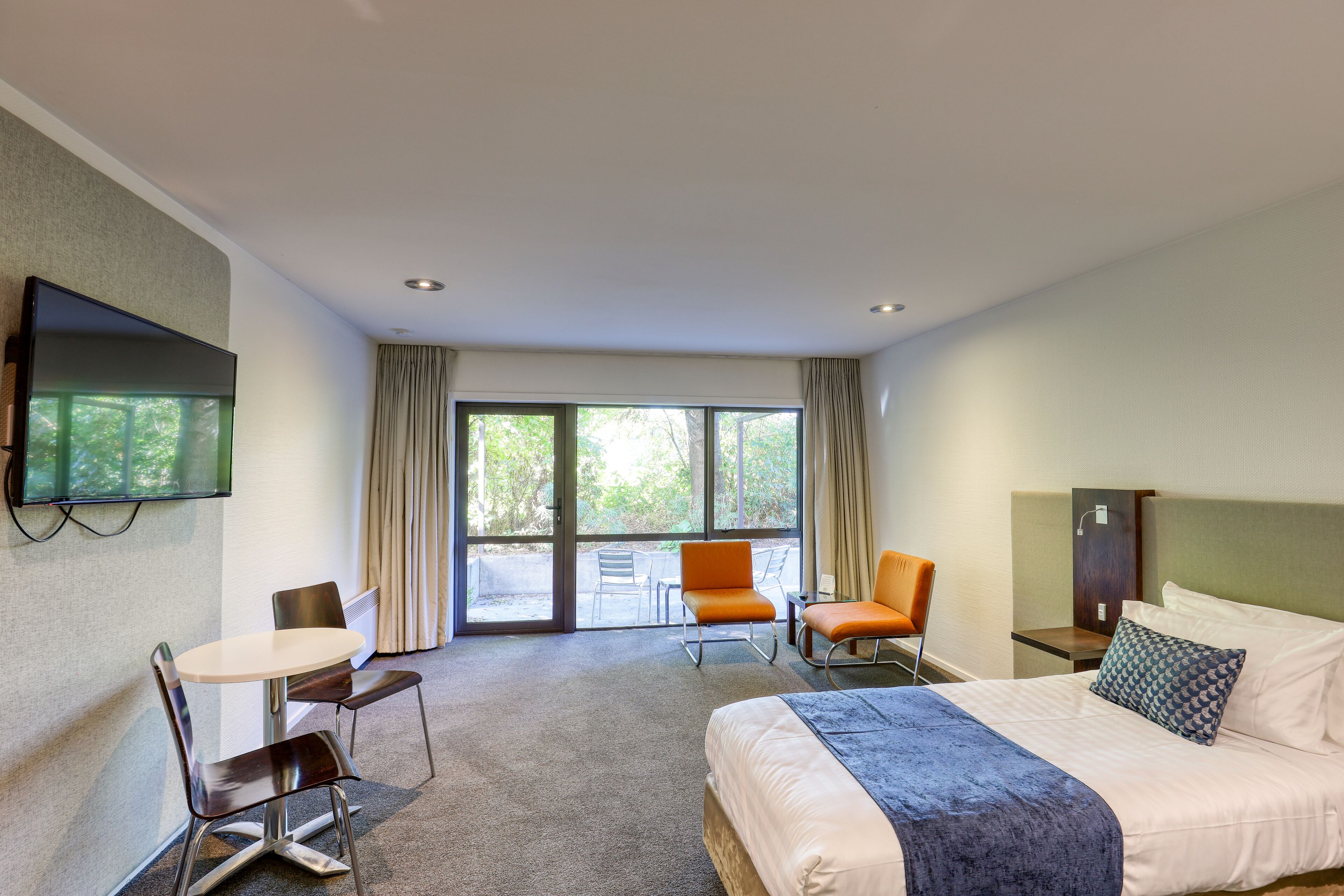 A hotel room with a bed, a desk, two chairs, a TV, and large windows.