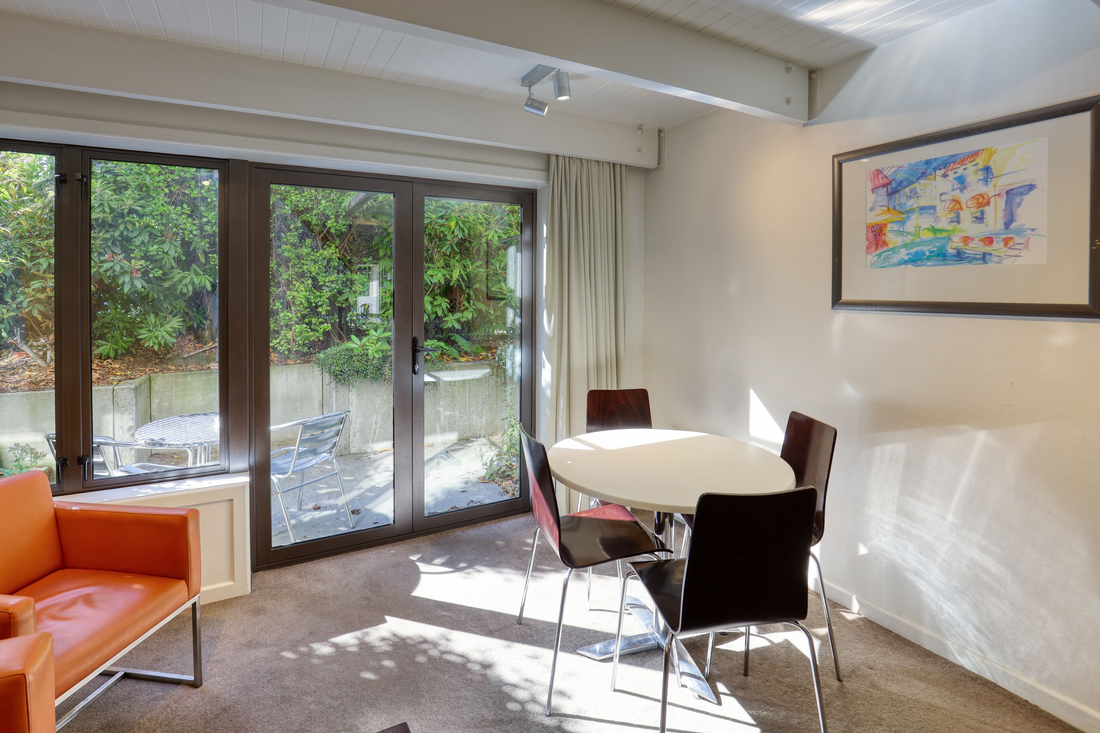 Een moderne kamer met een grote schuifdeur naar een tuin, een witte ronde tafel met vier stoelen, en een lijst met een abstract schilderij aan de muur.