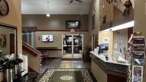 A hotel lobby with a reception desk, a coffee station, a TV mounted on the wall, and a patterned rug on the floor.