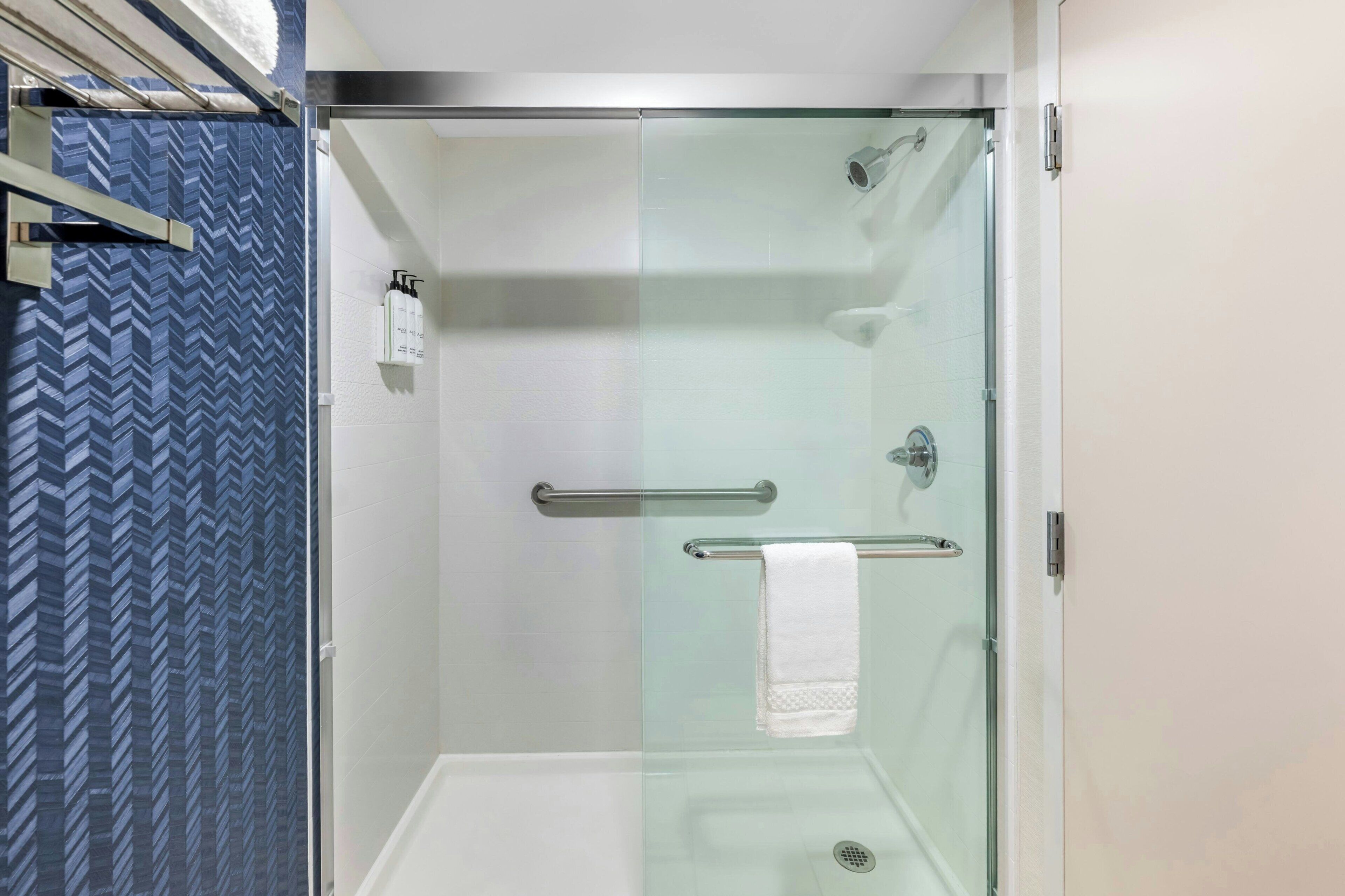 A glass-enclosed shower with a white towel hanging on a metal bar, a glass door, and a patterned wall to the left.