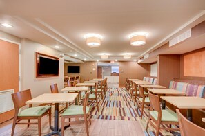 A hotel dining area with multiple tables and chairs, a television mounted on the wall, and a patterned carpet. - Fairfield Inn by Marriott Boston Woburn-Burlington (Woburn)