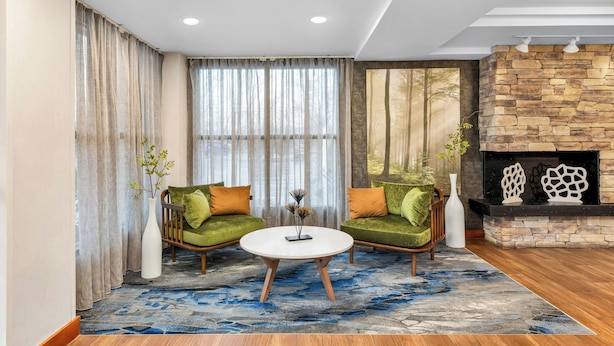 Un salon moderne avec deux canapés verts, une table basse ronde, une cheminée en pierre et de grandes fenêtres ornées de rideaux légers.