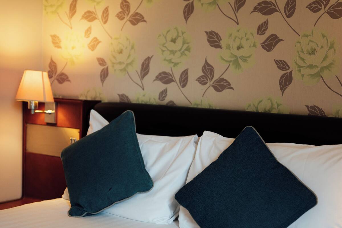 Una cama bien hecha con sábanas blancas y dos almohadas oscuras, una lámpara de noche y una pared con estampado floral.