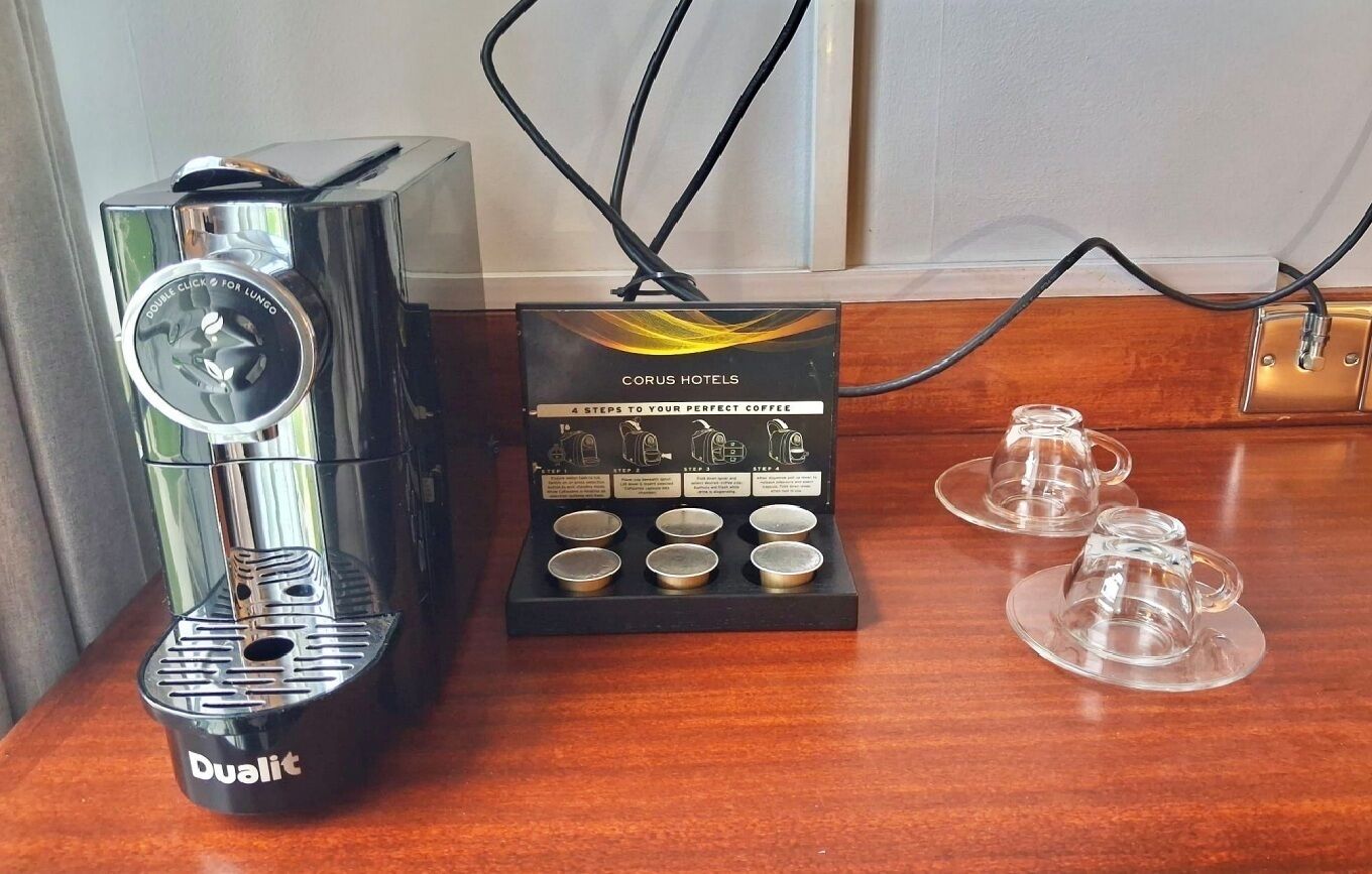A coffee machine with a display panel titled "CORUS HOTELS A STEPS TO YOUR PERFECT COFFEE" and a set of six cups on a wooden table.