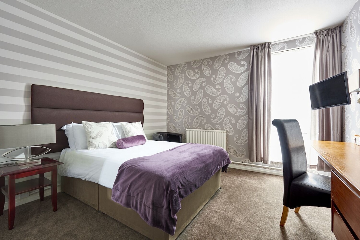 A hotel room with a bed, a desk, a chair, a TV, and a window with curtains.