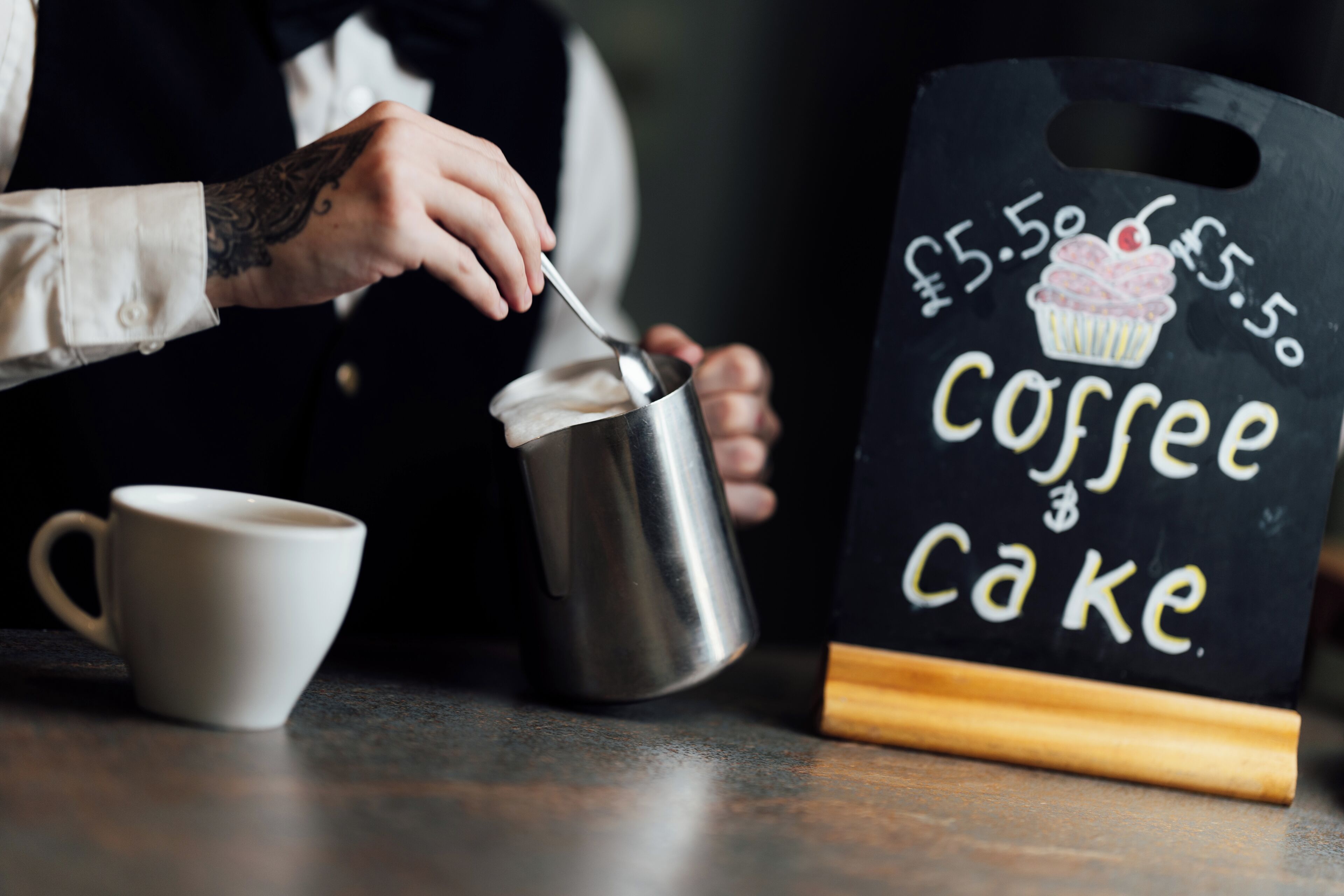 Una persona sirviendo una bebida en una taza, un letrero de pizarra con la leyenda 'Coffee Cake' y precios, y una pequeña taza blanca sobre una mesa.