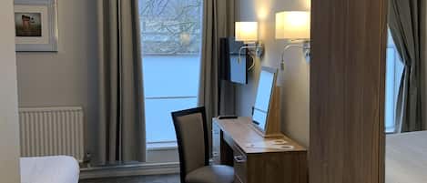 Habitación de hotel con cama, escritorio con computadora, silla, ventana con cortinas y radiador.