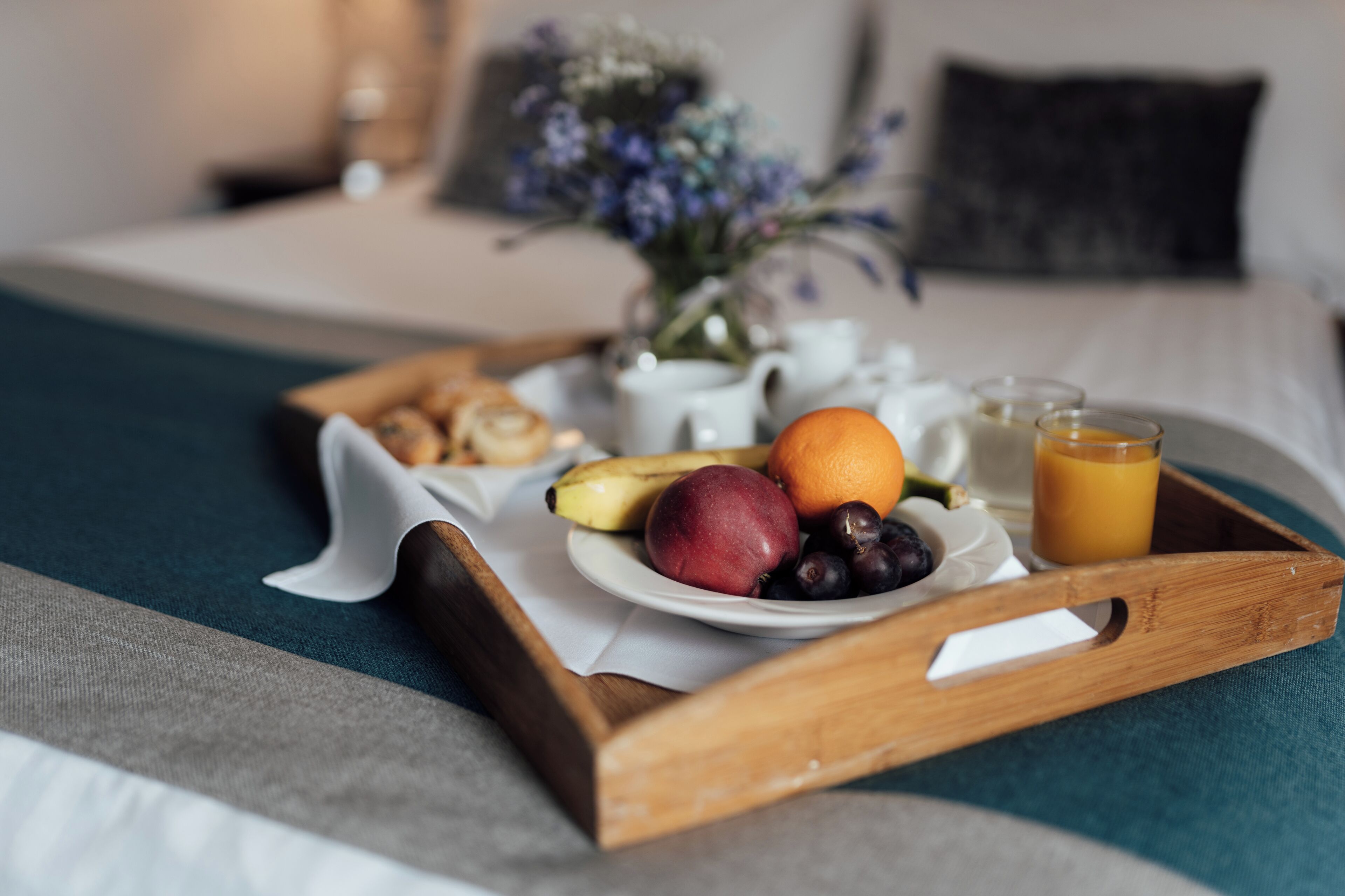 Un plateau de petit-déjeuner avec des fruits, un verre de jus d’orange et une théière blanche, posé sur un lit recouvert d’une couverture bleue et agrémenté d’un vase de fleurs.