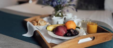 Bandeja de desayuno con frutas, un vaso de jugo de naranja y una tetera blanca sobre una cama con una manta azul y un florero con flores.