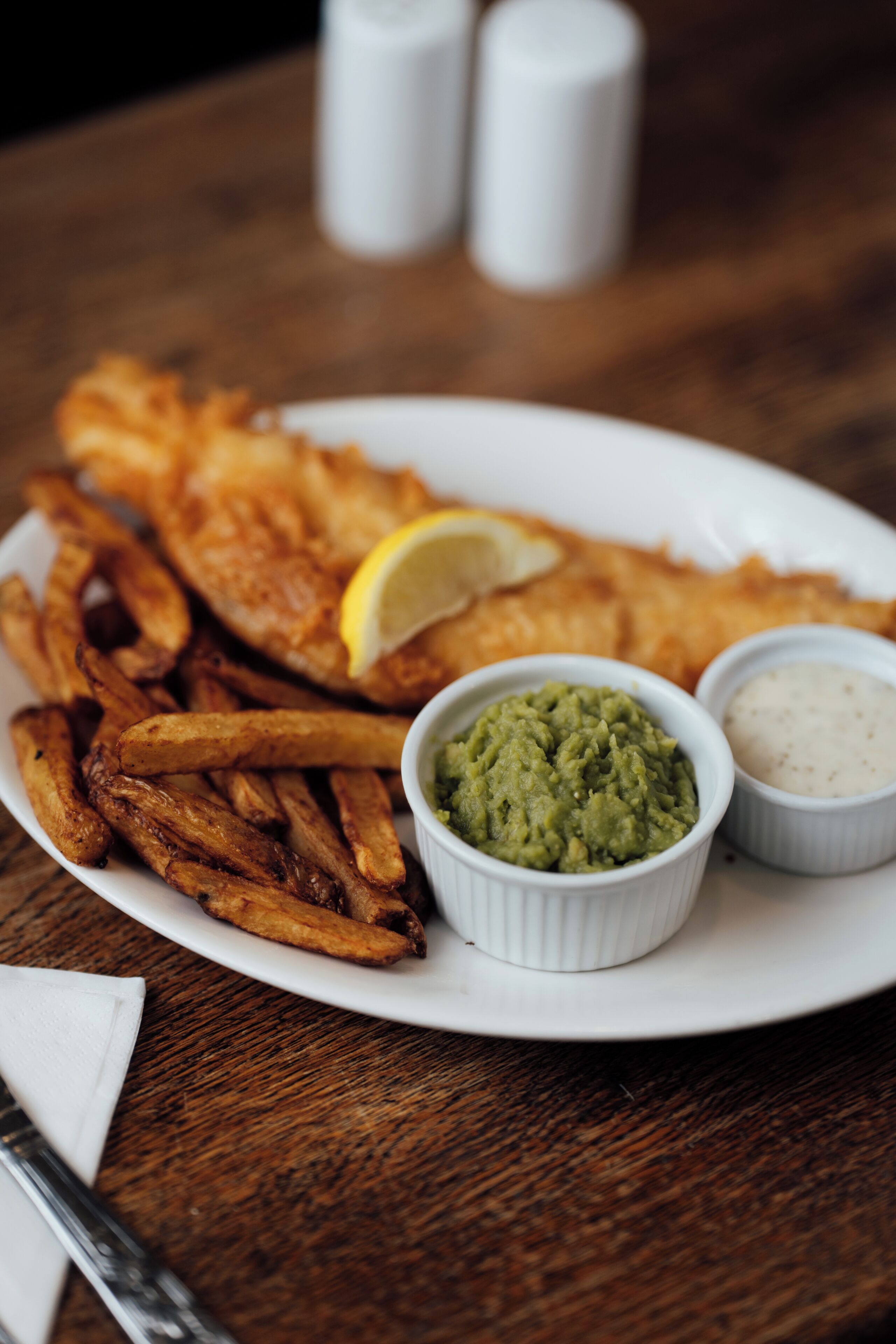 A plate with fish and chips, a lemon wedge, and two small containers of sauces.