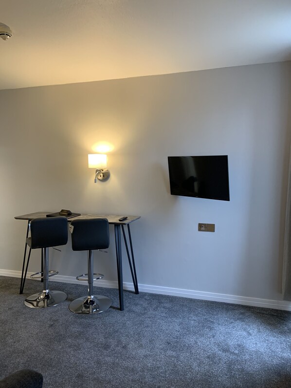 Una habitación pequeña y moderna con escritorio, dos sillas, televisor montado en la pared y una lámpara.