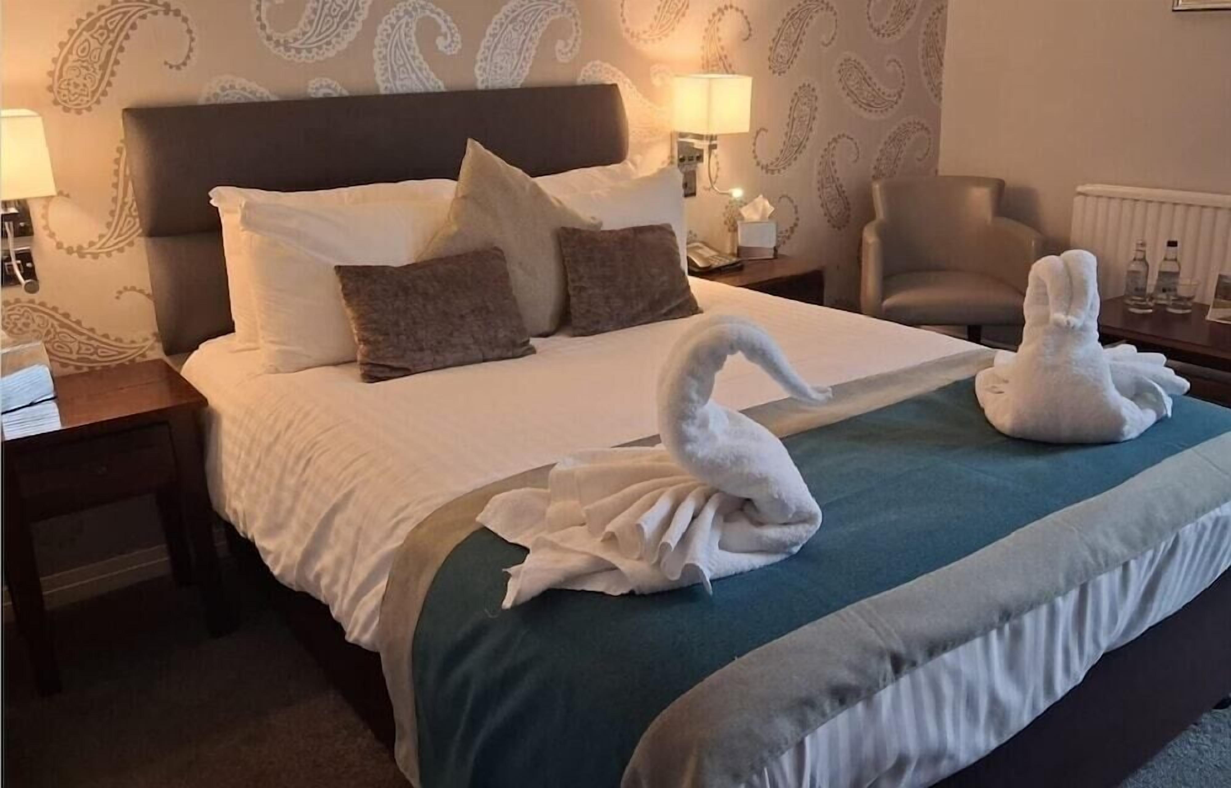 Quarto de hotel com cama bem arrumada, uma cadeira e uma luminária em uma mesinha de cabeceira.
