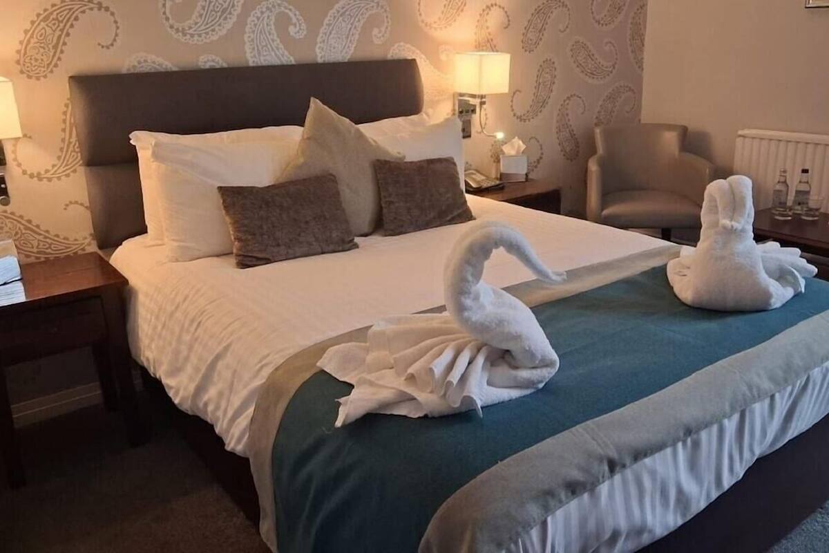 Una habitación de hotel con una cama bien hecha, una silla y una lámpara sobre una mesita de noche.