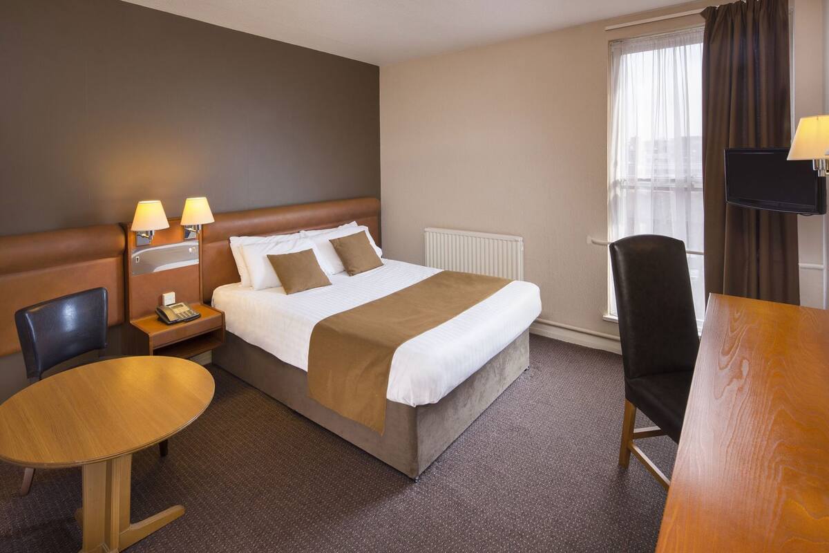Una habitación de hotel con cama, lámparas de noche, un escritorio, una silla y un televisor.