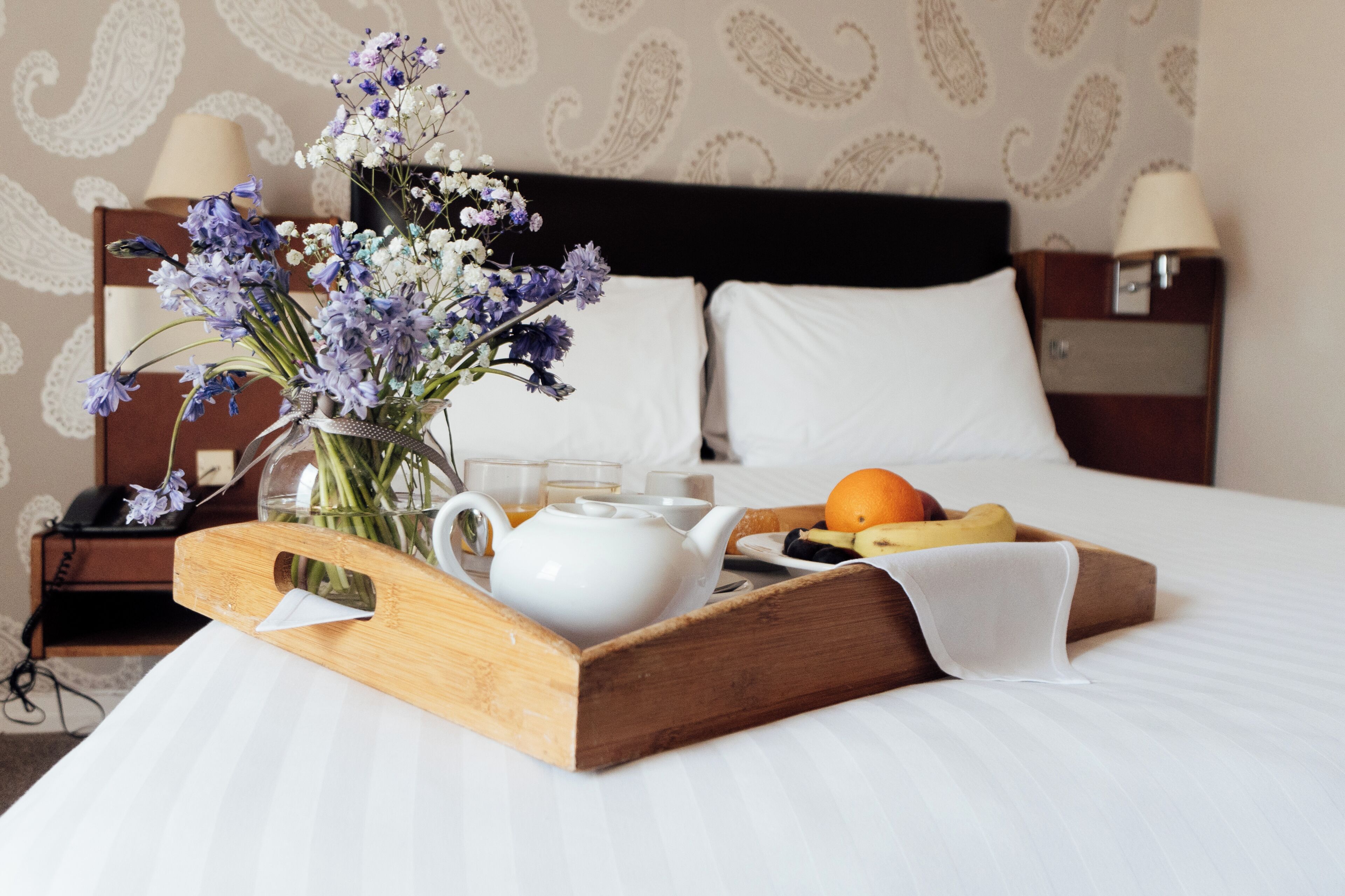 A neatly made bed with a tray of breakfast items, including a teapot, fruit, and flowers.