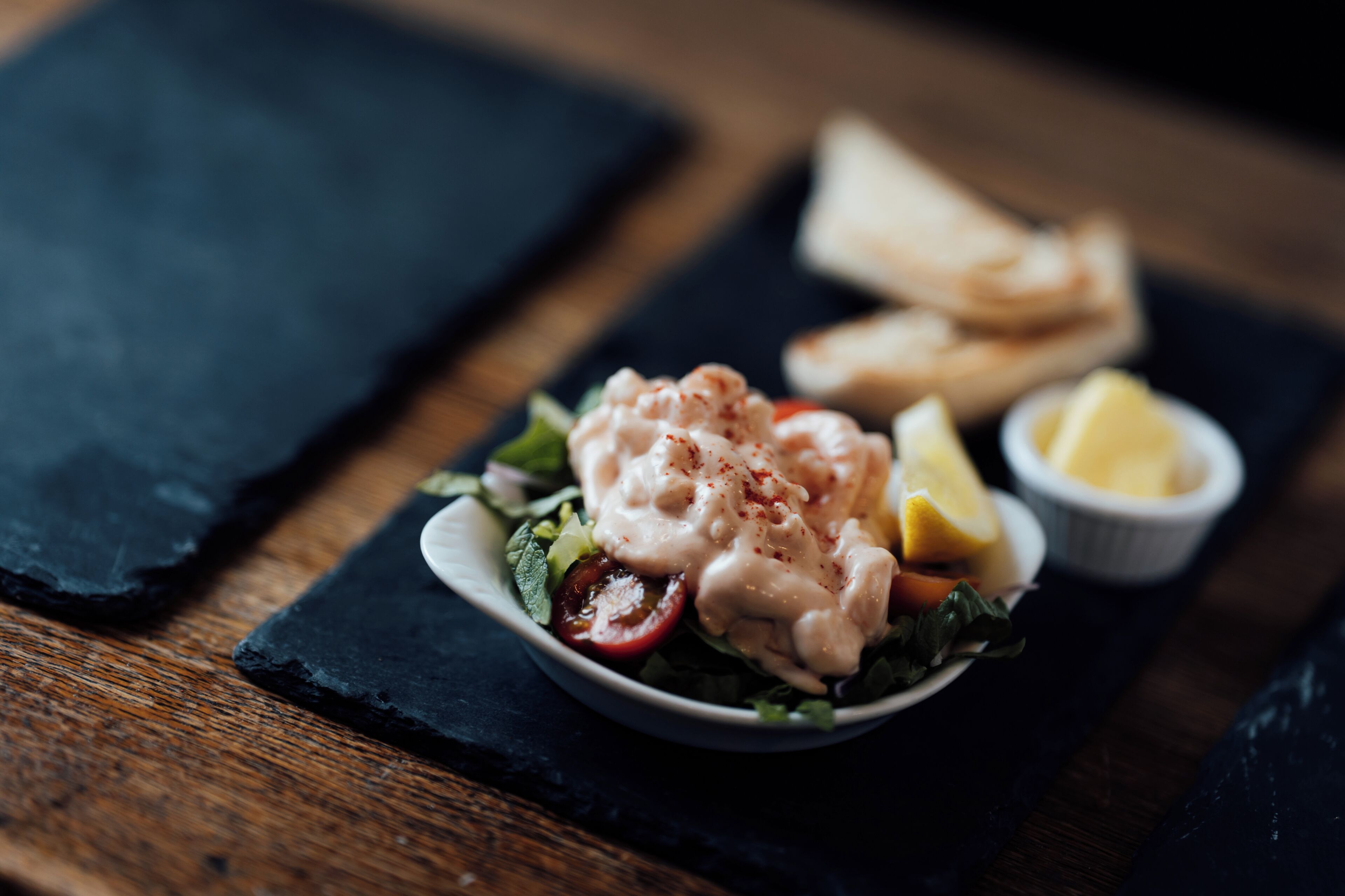 A plate of food with a creamy sauce, a lemon wedge, and a small bowl of sauce on a slate board.