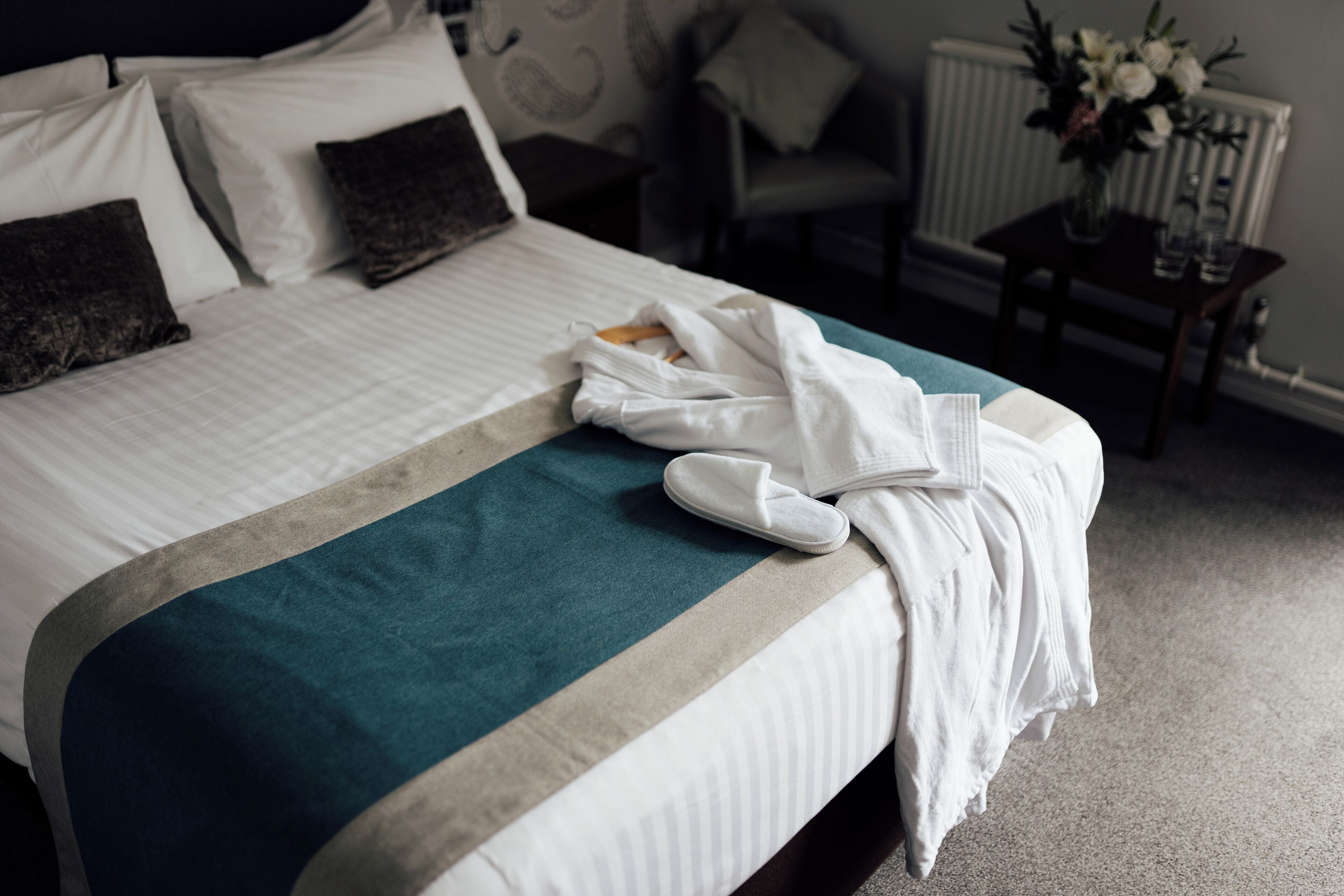 A neatly made bed with a teal and white bedspread, a white bathrobe, and slippers on it.