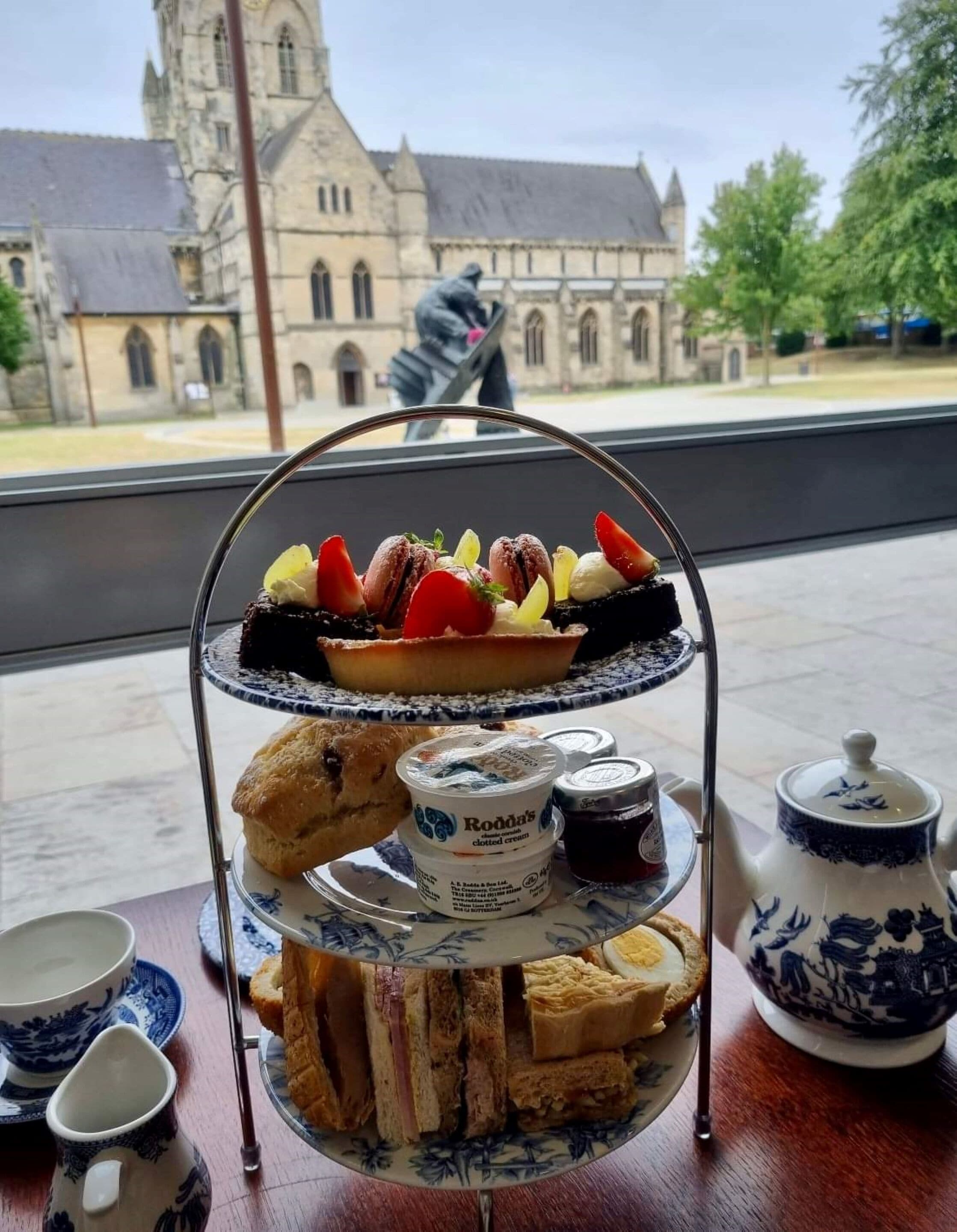 Eine gestufte Platte mit Speisen, darunter Gebäck, Sandwiches und Desserts, mit Blick auf ein historisches Gebäude im Hintergrund.