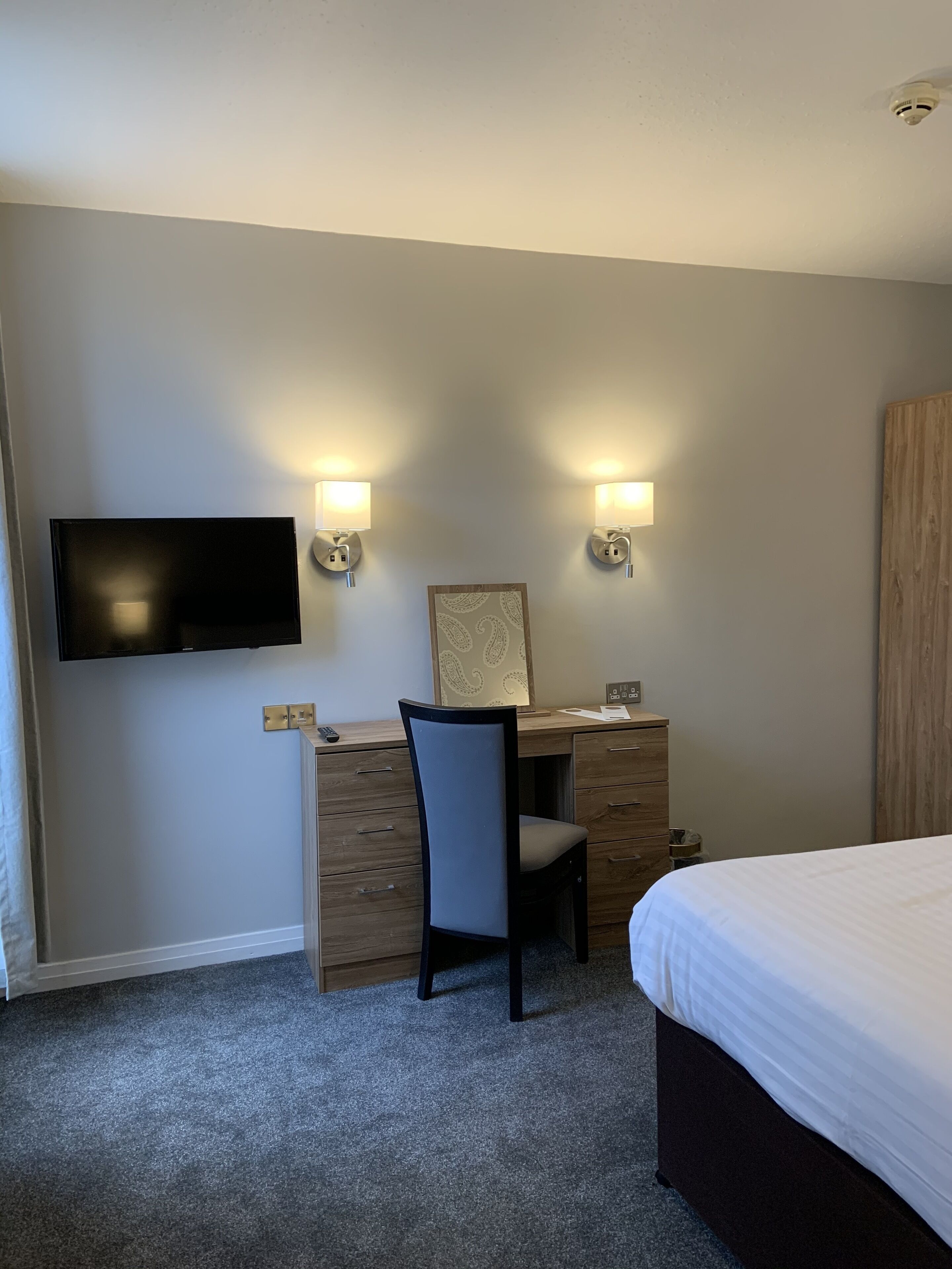A hotel room with a bed, a desk with a chair, a flat-screen TV mounted on the wall, and two wall-mounted lamps.