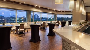 A modern dining area with round tables and chairs, large windows with a city view, and a long counter with glasses and a sink. - Westin Tampa Downtown Waterside (Tampa)