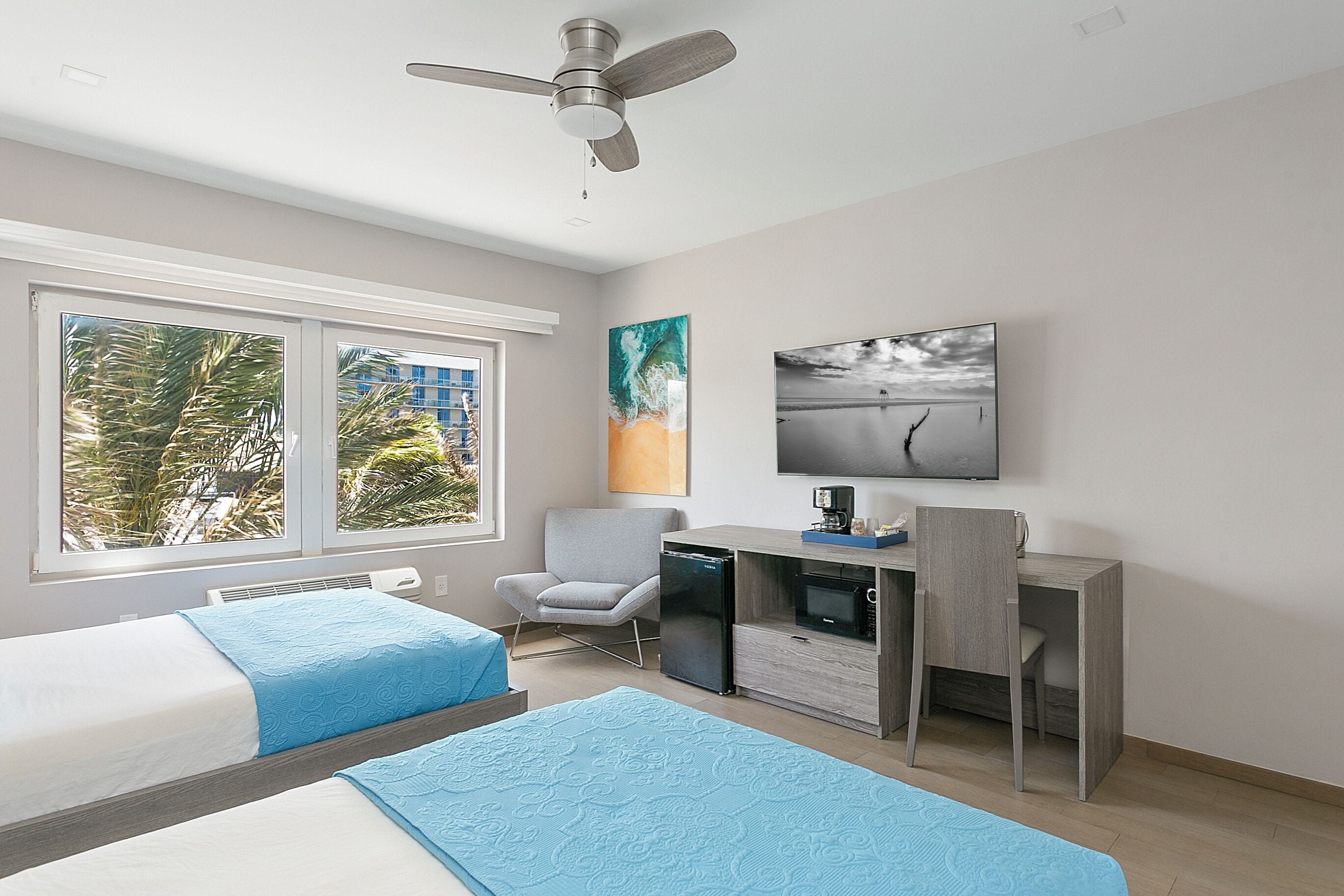 A hotel room with two beds, a desk with a microwave, a chair, a ceiling fan, and artwork on the walls.
