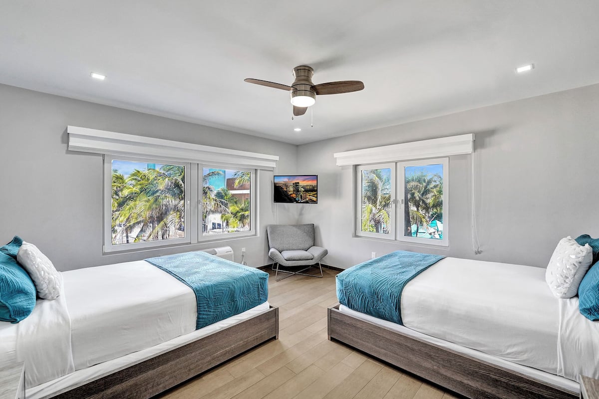 A bedroom with two beds, a ceiling fan, a TV, and large windows with tropical views.