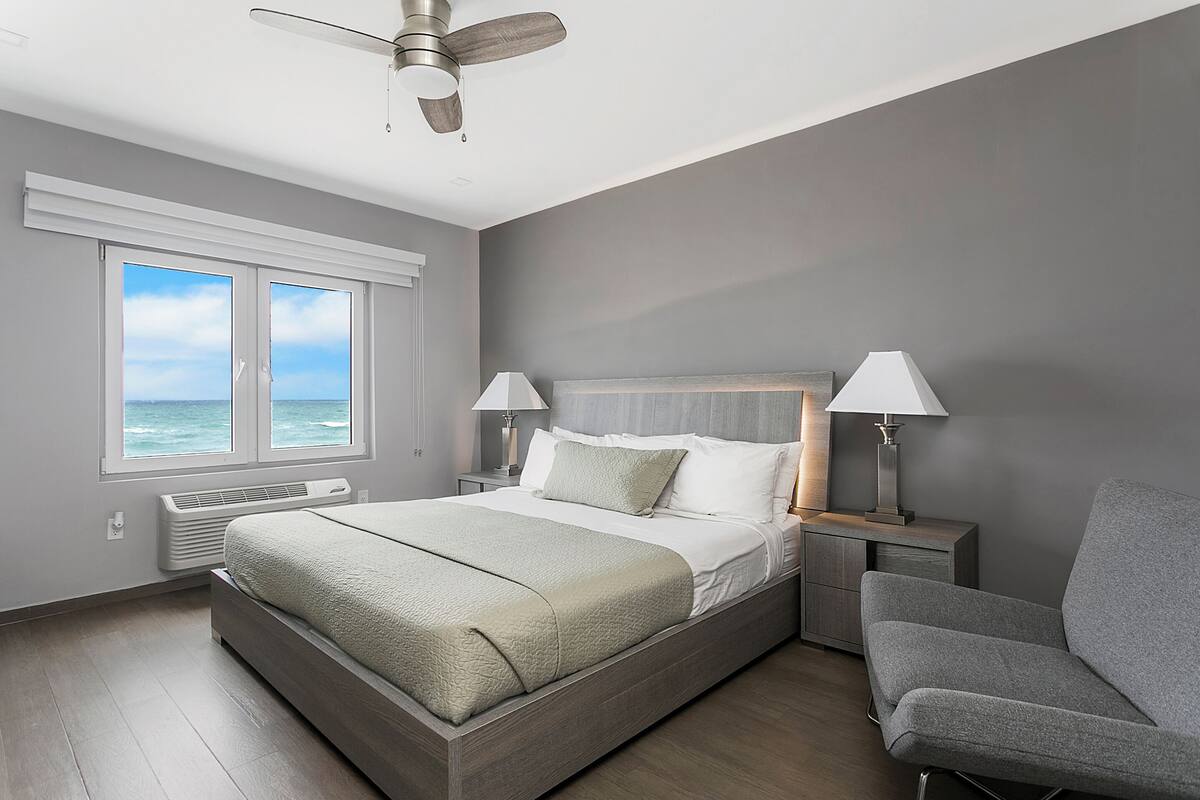 A modern bedroom with a large bed, two bedside lamps, a ceiling fan, and a view of the ocean through the windows.