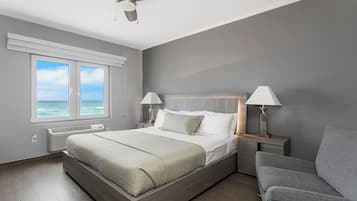 A modern bedroom with a large bed, two bedside lamps, a ceiling fan, and a view of the ocean through the windows.