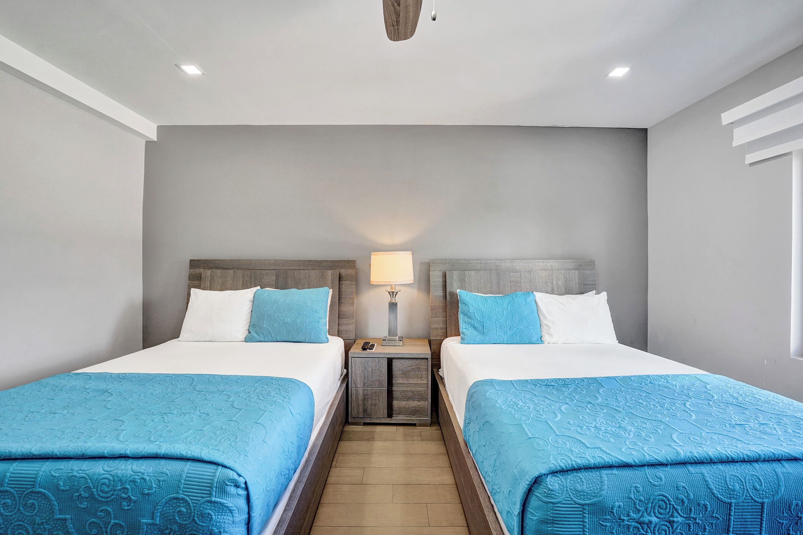 A hotel room with two beds, a nightstand with a lamp, and a window.