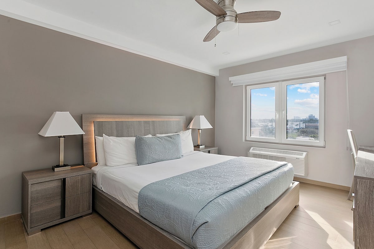 A modern bedroom with a large bed, two bedside tables with lamps, a ceiling fan, a radiator, and a window with a city view.