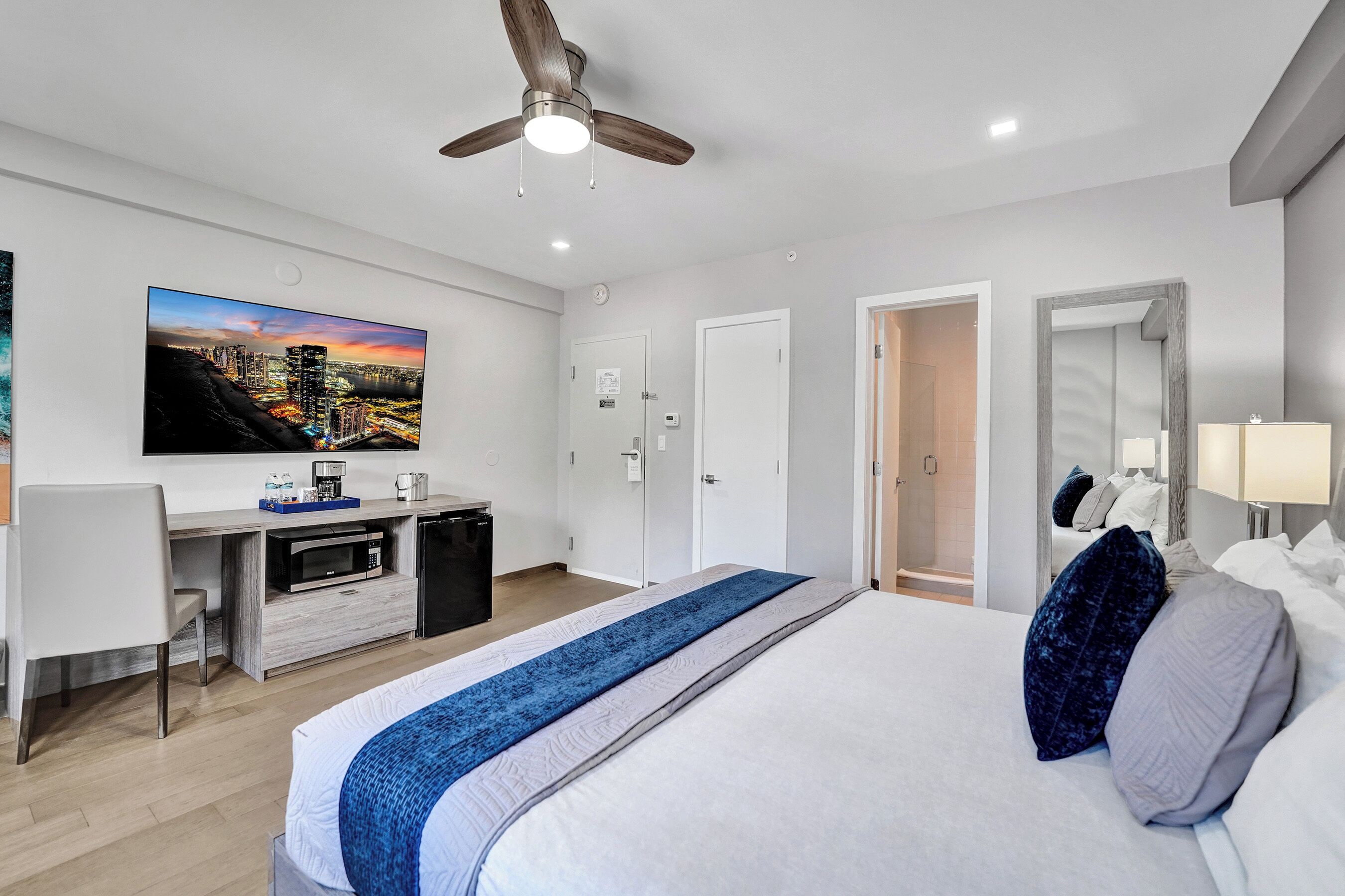 Una habitación de hotel moderna con una cama grande, un escritorio con televisión de pantalla plana, un ventilador de techo y un cuadro de un paisaje urbano en la pared.