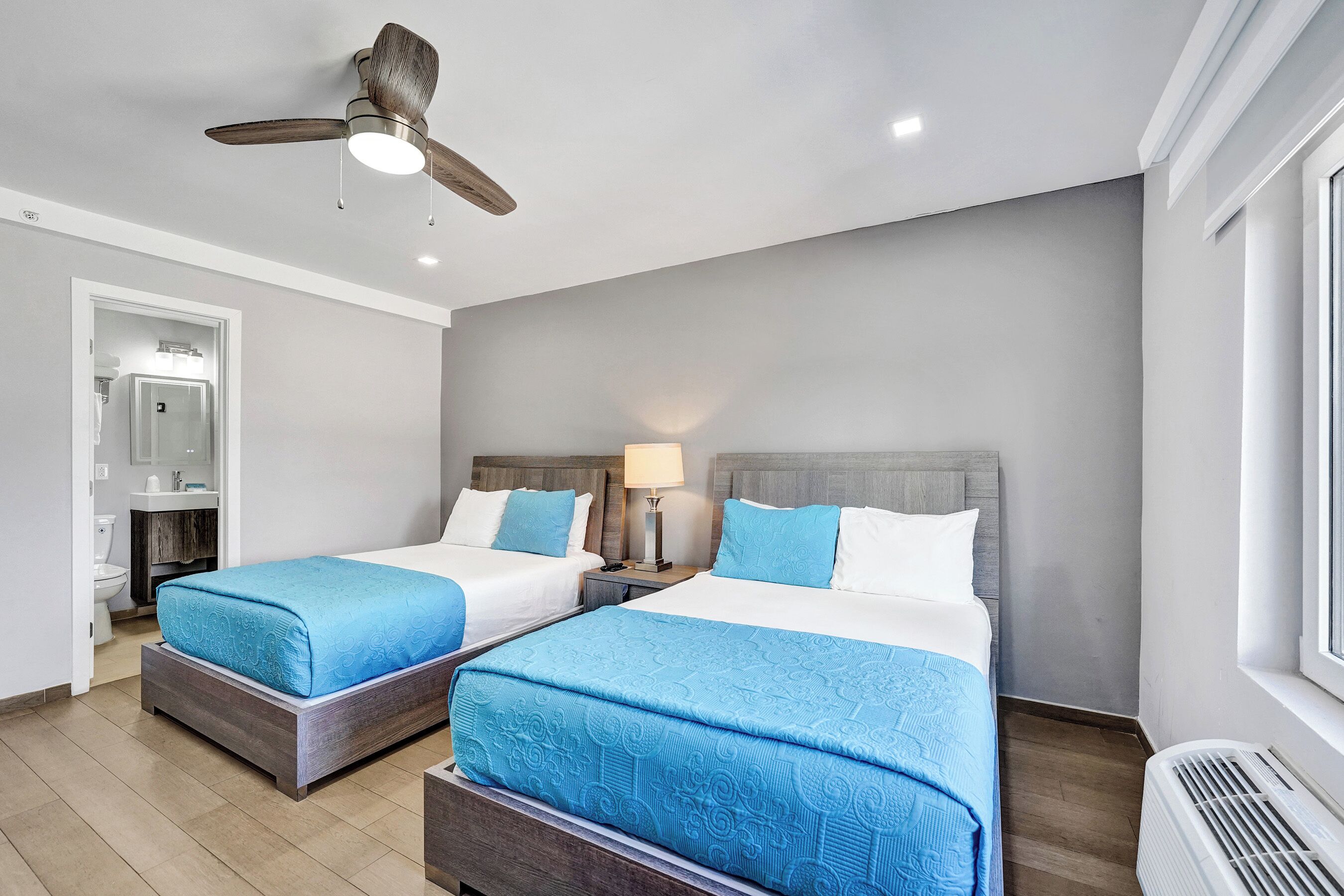 A hotel room with two beds, a ceiling fan, a window, and a bathroom visible through an open door.