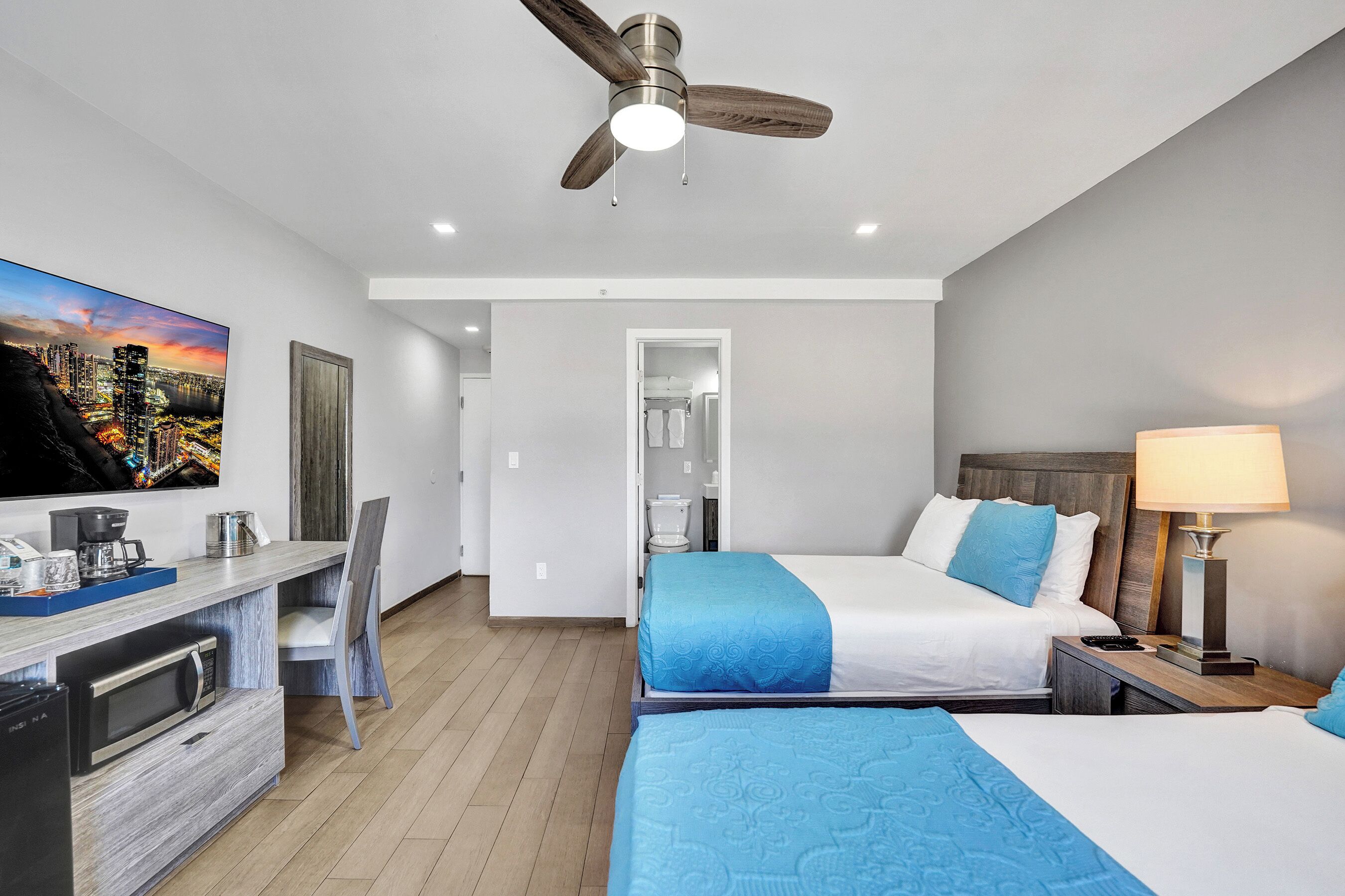 A hotel room with a bed, bedside table, lamp, TV, desk, chair, and a ceiling fan.