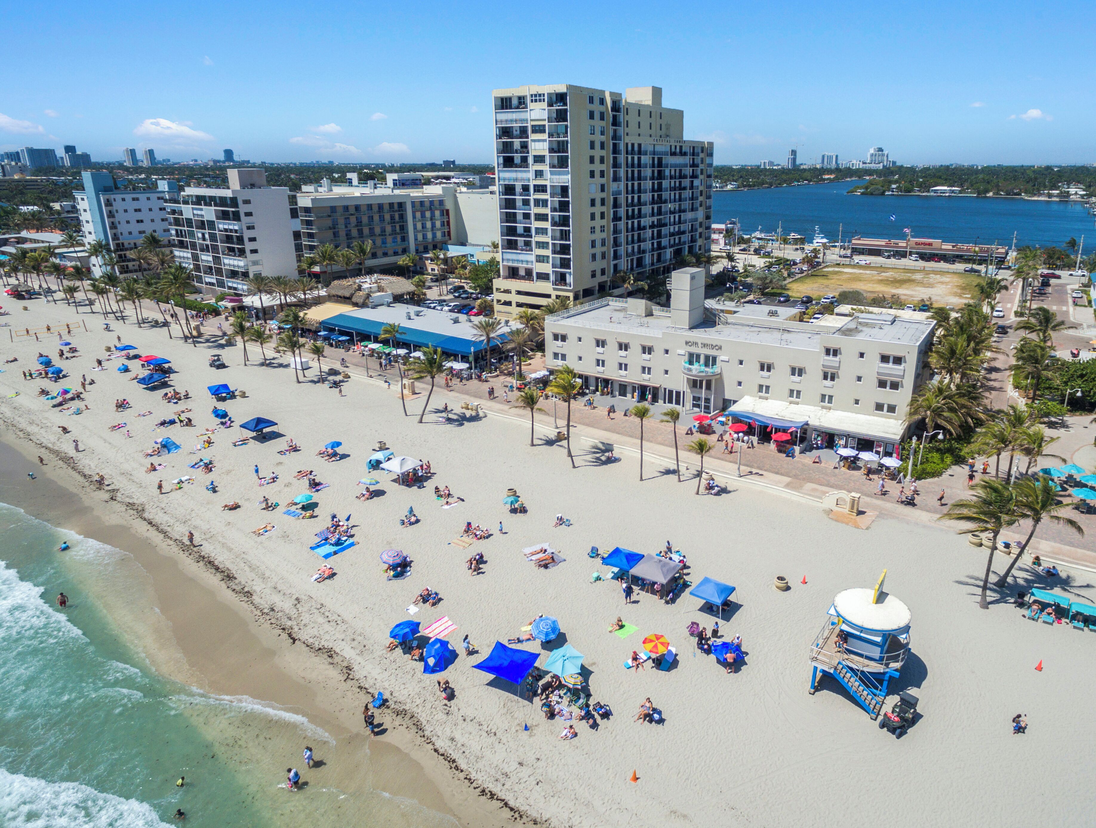 Un hôtel situé en bord de mer, avec plusieurs étages, un poste de surveillance pour les sauveteurs et une promenade en bord de plage agrémentée de parasols et fréquentée par les estivants.