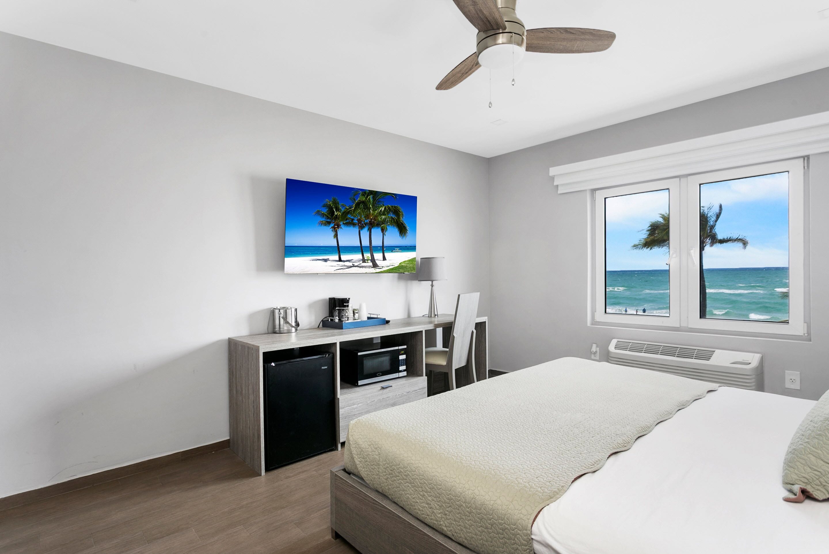 Een slaapkamer met een bed, bureau en stoel, een plafondventilator, een raam met uitzicht op de oceaan, en een schilderij van palmbomen en een strand.
