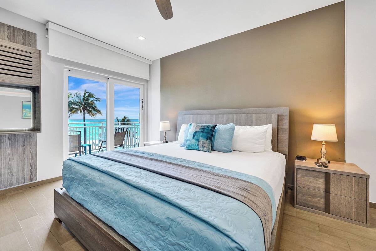 A bedroom with a large bed, a nightstand, a view of the ocean, and a balcony with a palm tree.