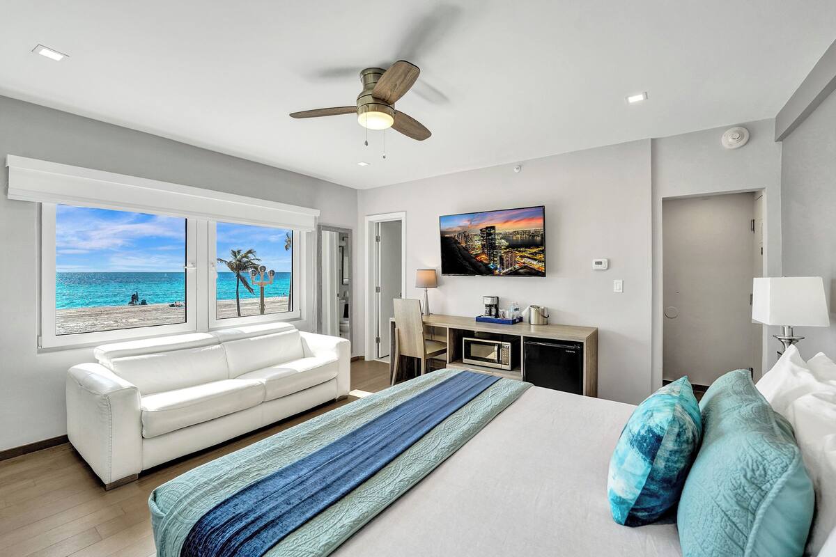 A hotel room with a bed, a white sofa, a desk with a computer, and a view of the beach.